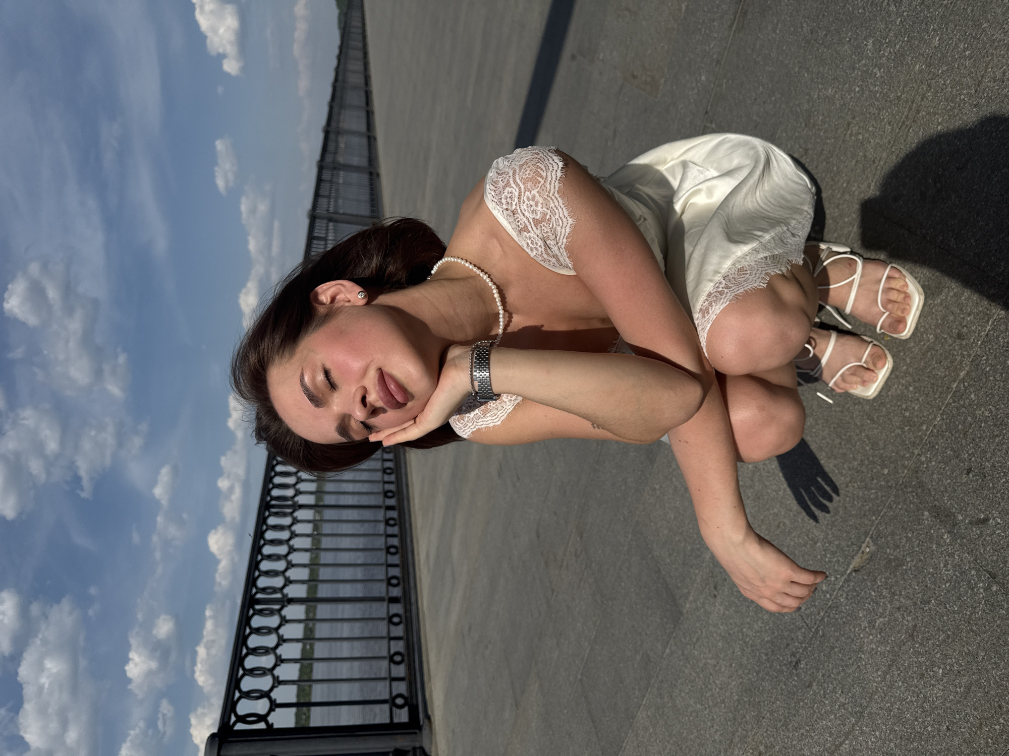A young woman is crouching on a gray paved surface, likely a promenade or sidewalk, with her left arm supporting her head. She is dressed in a white slip dress with lace details and white strappy sandals, also wearing a pearl necklace and a wristwatch. She faces the camera with her eyes closed and tongue slightly sticking out. To the left of the woman, a black wrought-iron railing runs along what appears to be a river, and beyond the railing, the sky is visible with white clouds. The location is Perm, Russia.