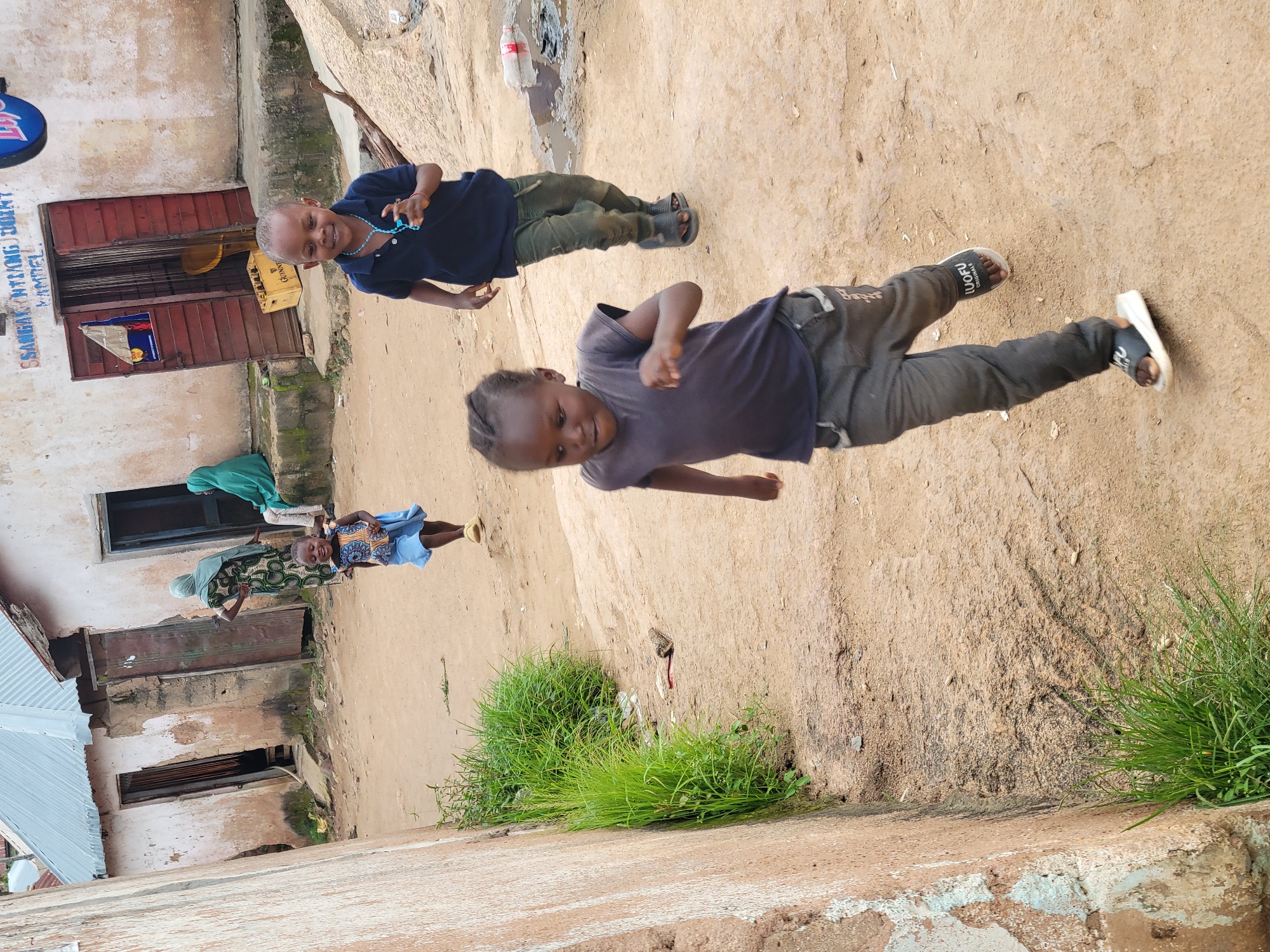 This image captures a candid moment in a street setting in Jos, Nigeria, likely on a bright, overcast day. The foreground is dominated by a young Black child, appearing to be a girl, running energetically towards the viewer. She is wearing a dark grey t-shirt and dark grey pants. Her expression is one of joy and exertion, with her mouth open and eyes focused ahead. Behind her, slightly to the right and further back, another young Black child, presumably a boy, is also running in the same direction. He is wearing a dark blue polo shirt, dark green pants, and dark sandals. He is looking towards the viewer and has a playful smile. Further in the background, another child in a blue dress is running away from the viewer. Two women in headscarves are visible near some buildings. One woman in a green headscarf is standing and looking towards the children, while another woman in a green headscarf is seated. The environment is a dusty, unpaved street with sparse vegetation growing along the side of a textured, light-colored wall on the left. In the background, there are simple, weathered buildings with corrugated metal roofs and wooden doors, suggesting a residential or mixed-use area. A sign with text, including "SSANGANTIANG DOINT", is visible above a doorway. A crate of Guinness beer is visible near one of the buildings, and a discarded plastic bottle lies in a puddle of water to the right. The overall scene depicts children engaged in play in a typical urban street environment.