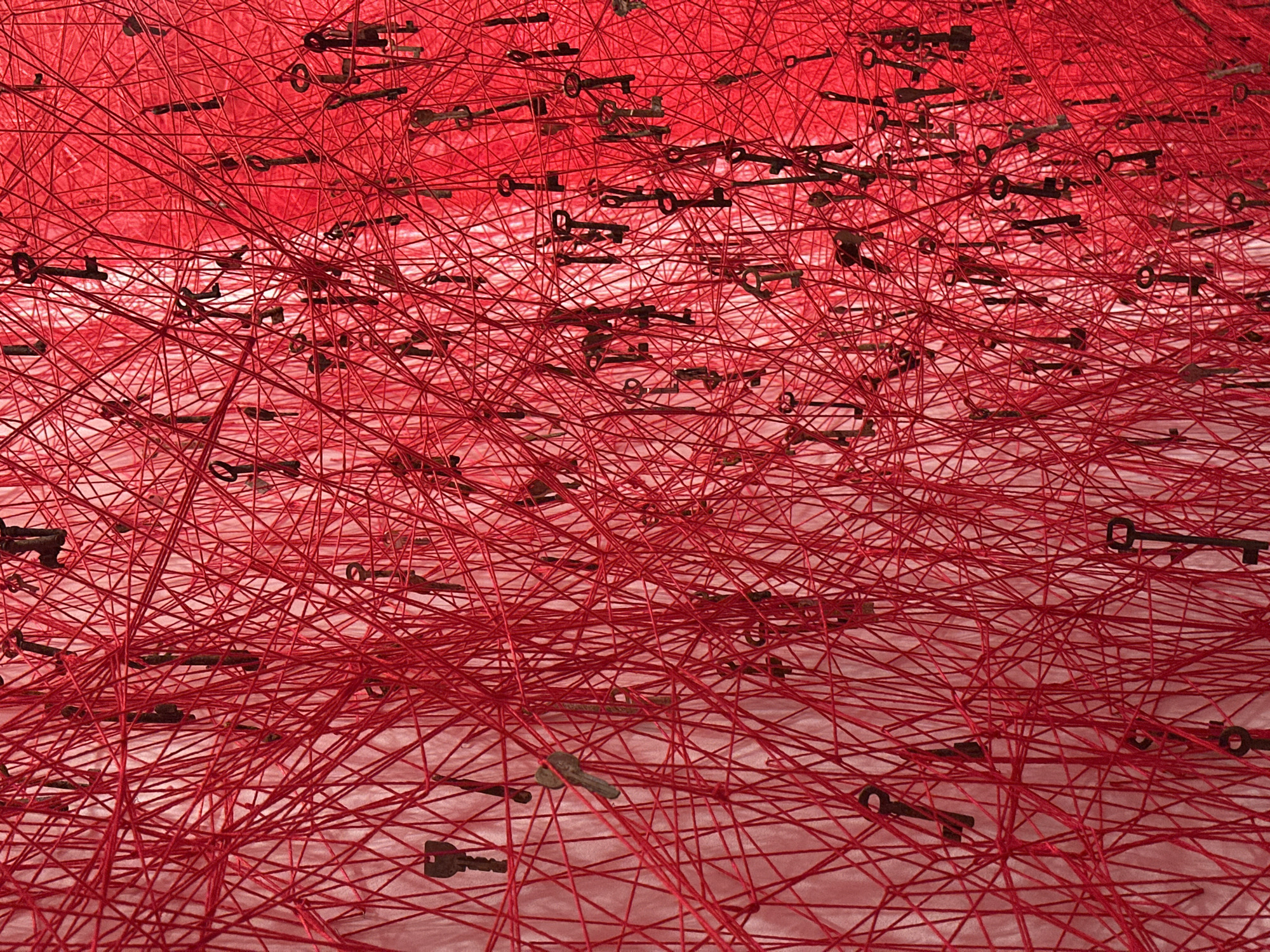 The image depicts an art installation featuring a dense network of red threads suspended in space, with antique-looking keys attached at various points along the threads. The background appears to be a pale pink or white, possibly the floor or a wall. The keys are predominantly a dark, aged metal color. The scene is likely an interior space due to the controlled lighting and apparent absence of natural elements. While a specific location within London is not visually discernible from the image, this type of installation could be found in a gallery or museum setting within the city.