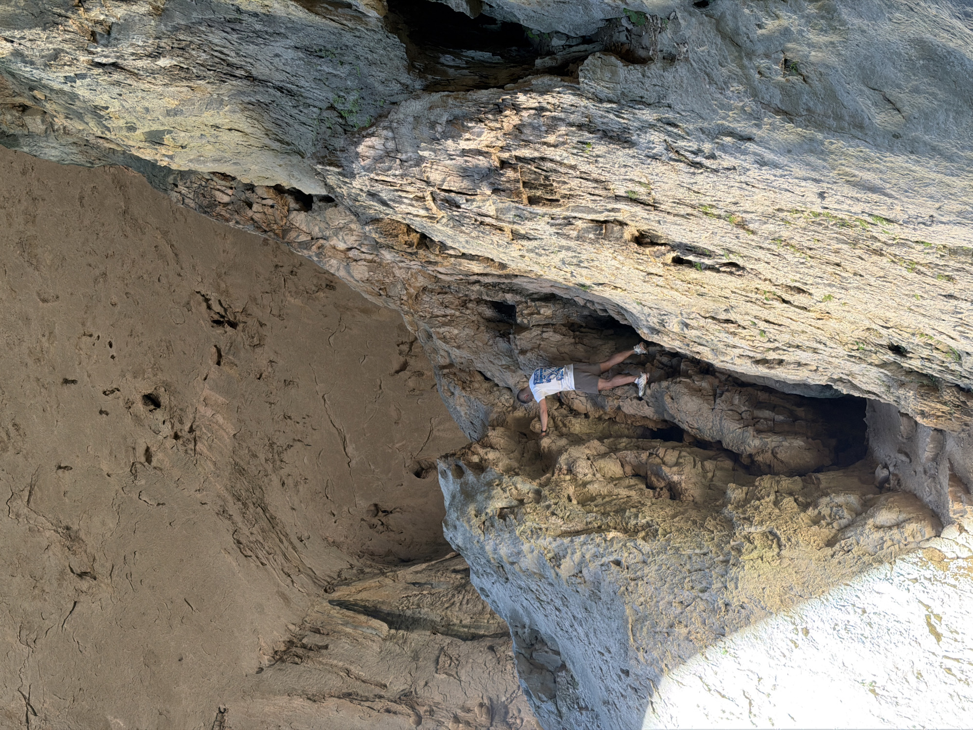 The image shows a man rock climbing in Devetashka Cave, near the village of Karlukovo, Bulgaria. The cave walls are textured and gray. The man is wearing a white t-shirt, khaki shorts, and sneakers. He is positioned horizontally across a rocky outcrop. The background includes other parts of the cave, with sunlight visible in the lower right corner.