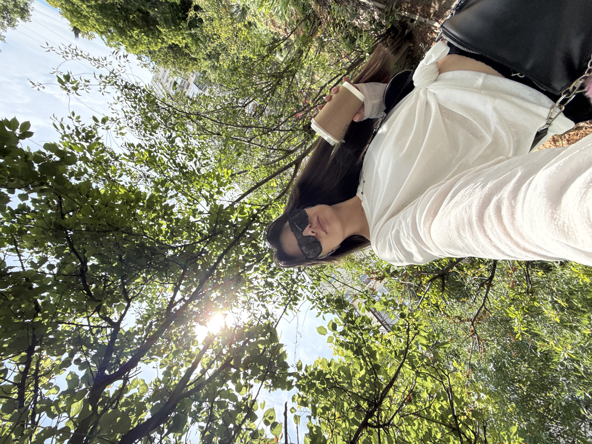 A woman is captured in a low-angle, selfie-style shot, holding a disposable coffee cup in her right hand. She is wearing a white, long-sleeved, knotted shirt and dark sunglasses. The background is dominated by green foliage and branches of trees, with dappled sunlight filtering through the leaves, creating bright spots and casting shadows. The sky is visible in patches, appearing bright blue with some white clouds. In the distance, a building with windows can be seen through the trees. The lower portion of the image shows a black purse with a silver chain strap. The ground appears to be covered with dry leaves and dirt. The overall setting suggests an outdoor urban environment, possibly a park or a tree-lined street, given the presence of the building.