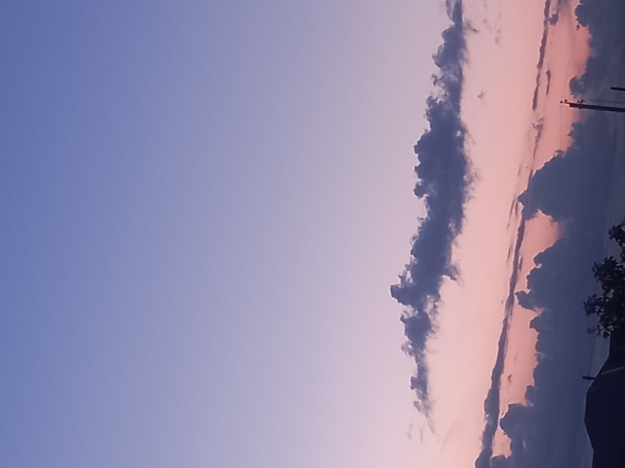 The image depicts a twilight sky over a rural landscape in Қозыкеткен ауылдық округі, Kazakhstan. The sky transitions from a light purple hue at the top to softer pinks and purples near the horizon, indicating either sunrise or sunset. Several dark, cloud formations are visible against the colorful sky. In the foreground, the silhouette of a building roof and some trees can be seen, suggesting a residential or agricultural area. A utility pole with wires is also present, further confirming a populated or developed area. The overall scene is tranquil and atmospheric, characteristic of a rural evening or early morning.