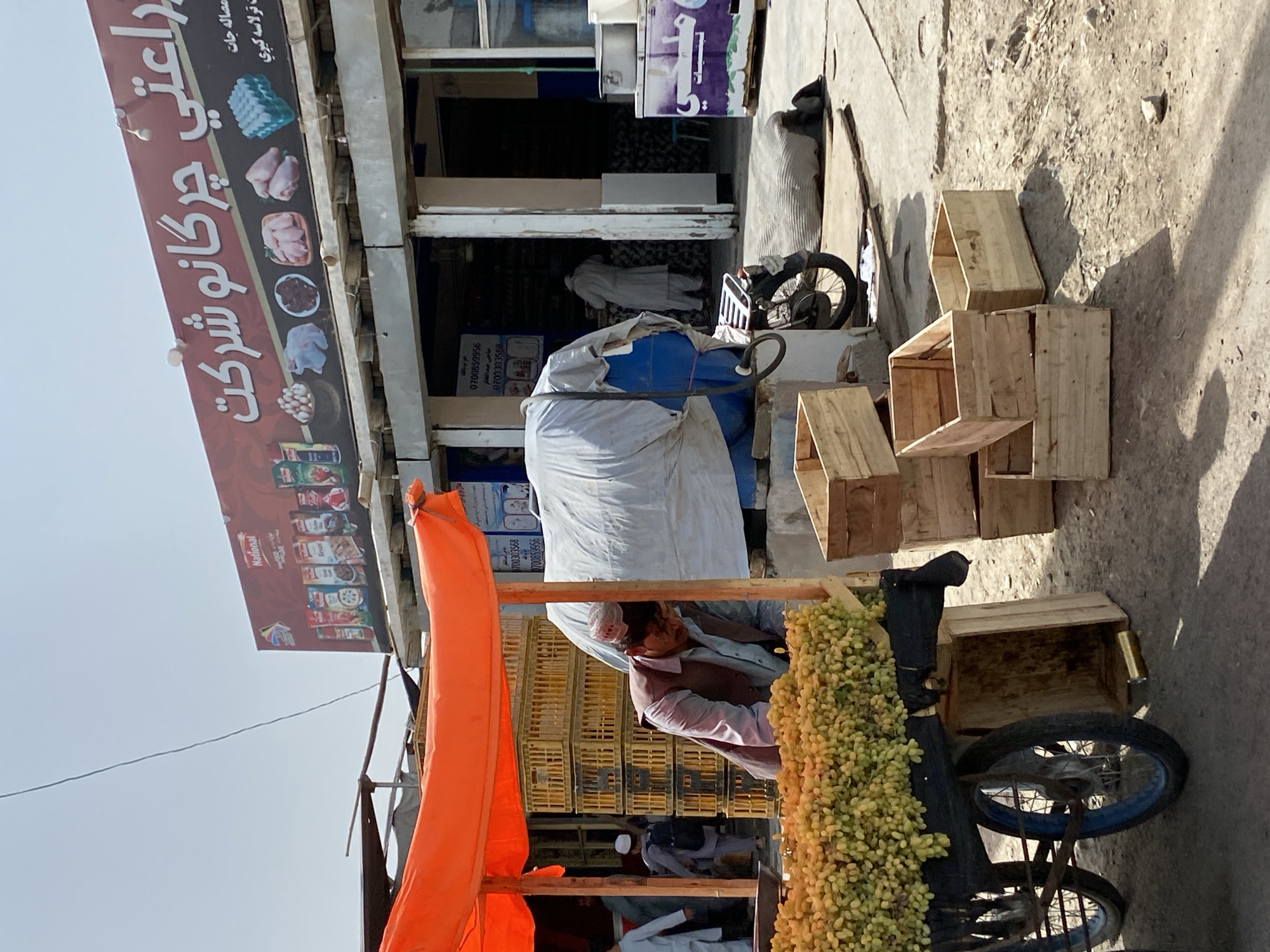In the city of Kandahar, Afghanistan, a street vendor stands behind a cart laden with bunches of light green grapes, his head bowed as he attends to his produce. The cart, made of metal with blue-rimmed wheels, is partially covered by an orange tarp providing shade. Behind the vendor, a stall is set up with stacked yellow crates. A large blue tarpaulin is draped over an unseen object to the right of the stall. Further back, the entrance to a shop is visible, with a prominent sign bearing Arabic script and images of food products. Several people are seen in the background: one person in traditional clothing walks away from the camera, another is partially visible near the shop entrance, and a third individual in a white outfit stands inside the shop. A motorcycle is parked near the shop. The ground is dusty and uneven, typical of an outdoor market. Wooden crates are stacked around the cart and in front of the shop. The sky is clear and bright.