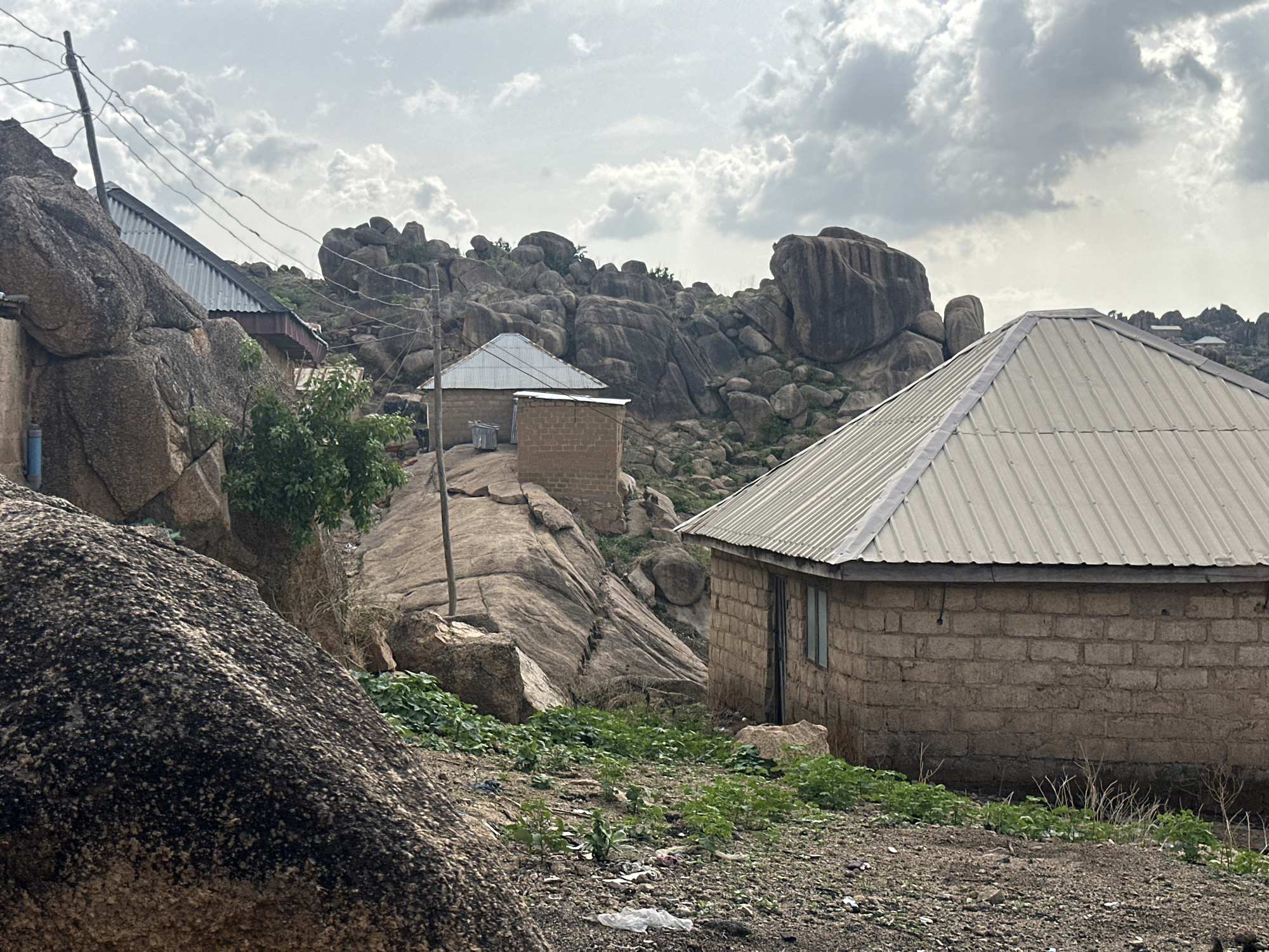 The image depicts a residential area in Jos, Nigeria, characterized by the presence of large rock formations interspersed with houses. The houses are constructed of cinder blocks and have corrugated metal roofs. The foreground features a large, dark-colored boulder, with vegetation and scattered debris on the ground. Power lines are visible in the background. The sky is overcast with grey and white clouds. The location is a residential area built among the rocky hills surrounding the city of Jos.