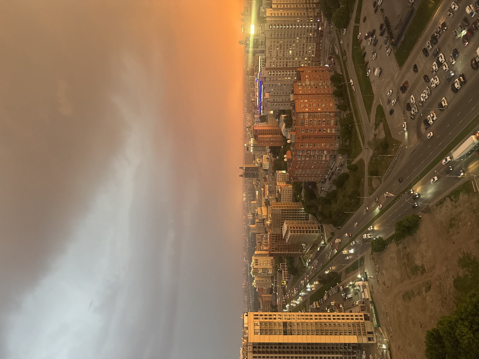 This is an aerial, long-shot photograph of a city skyline at dusk, likely in Novosibirsk, Russia, given the signage. The sky is dramatic, with a gradient of colors from a grey, overcast tone on the left to a vibrant, fiery orange and yellow on the right, suggesting a sunset or significant atmospheric phenomenon. The city below is densely populated with buildings of varying heights and architectural styles, many of them modern apartment complexes with illuminated windows.
A major highway runs diagonally through the scene from the lower left towards the center, populated with numerous vehicles, their headlights and taillights creating streaks of light on the wet road surface, indicating recent rain. Several parked cars are visible in parking lots adjacent to the buildings and along the roadside. Construction sites with bare ground and unfinished buildings are also present, particularly in the foreground on the left, featuring a tall, modern, beige-colored residential building under construction. One of these construction sites has a sign with Cyrillic text, "САКУРА ПАРК," which translates to "Sakura Park," a development name.
The overall impression is one of a bustling urban environment under a striking and dramatic sky, capturing a moment of transition between day and night, potentially after a rain shower. No individuals are clearly discernible in the image.