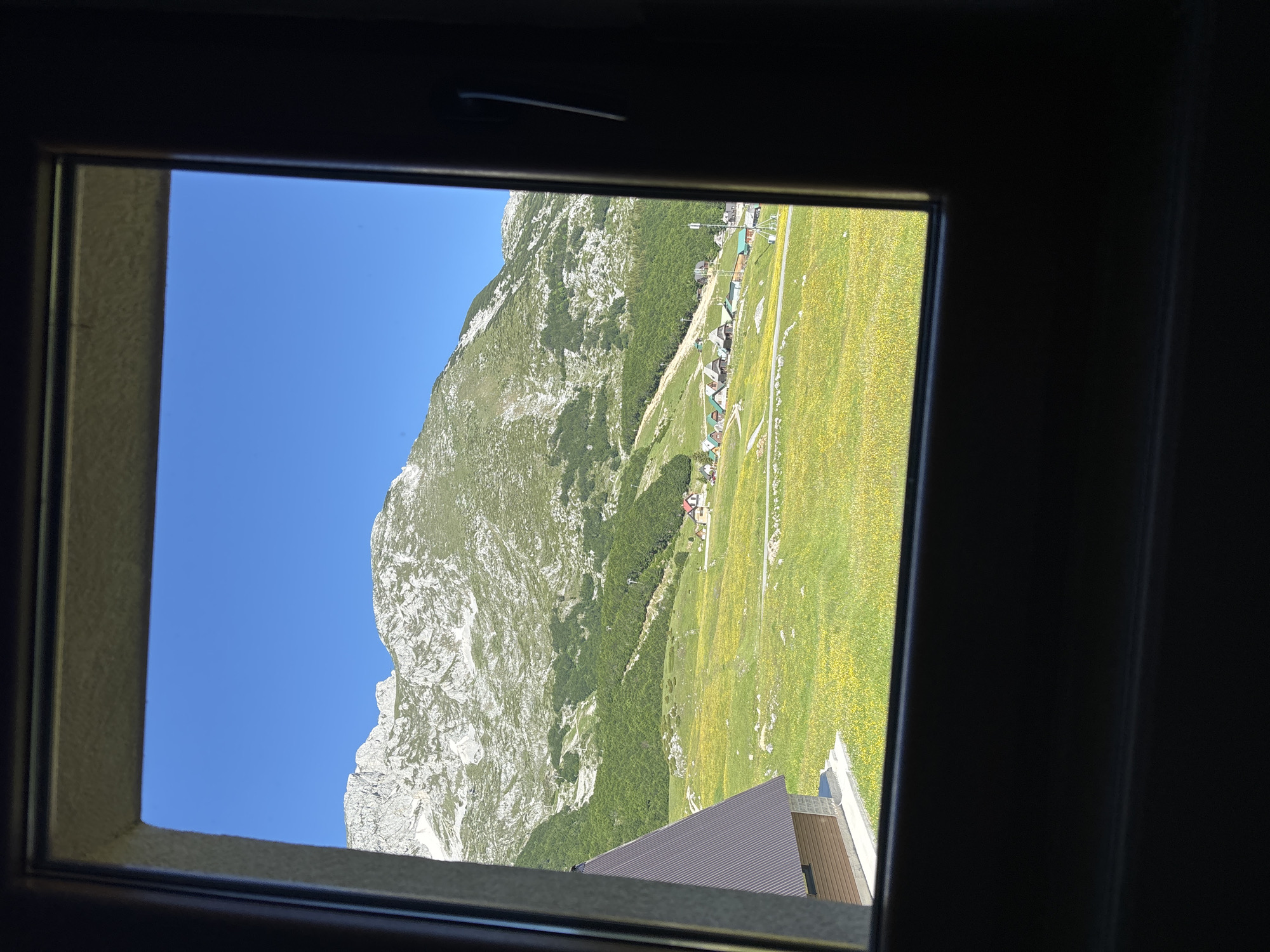 The image shows a view from a window in the city of Virak, Montenegro, looking out onto a mountain landscape. In the foreground, to the left, is a brown, angular building with a dark brown roof, partially framed by the window. Beyond this building, a vast, green meadow is dotted with yellow wildflowers. A paved path winds through the meadow, leading towards a cluster of small, colorful buildings with green roofs, appearing to be houses or cabins. Further in the distance, a large, verdant mountain rises sharply, its upper slopes covered in light-colored, rocky terrain. The sky above is a clear, bright blue. The window frame, dark and somewhat blurred, encloses the entire scene.