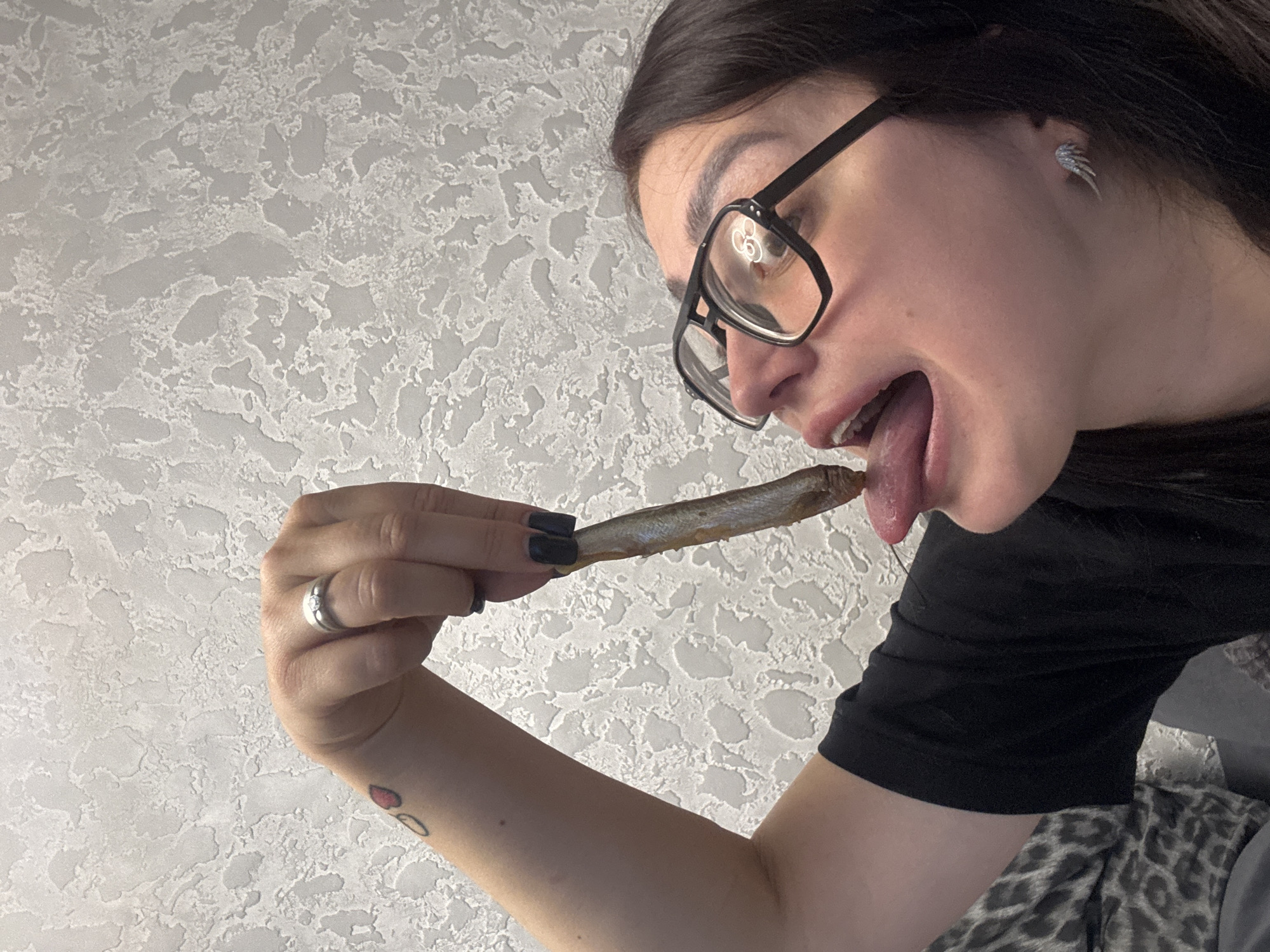 A woman in a black shirt and glasses is holding a small fish with her right hand and licking it with her tongue. She has black nail polish and a ring on her right ring finger. There is a small tattoo of a heart with an arrow through it on her left arm. The background is a textured, light-colored wall.