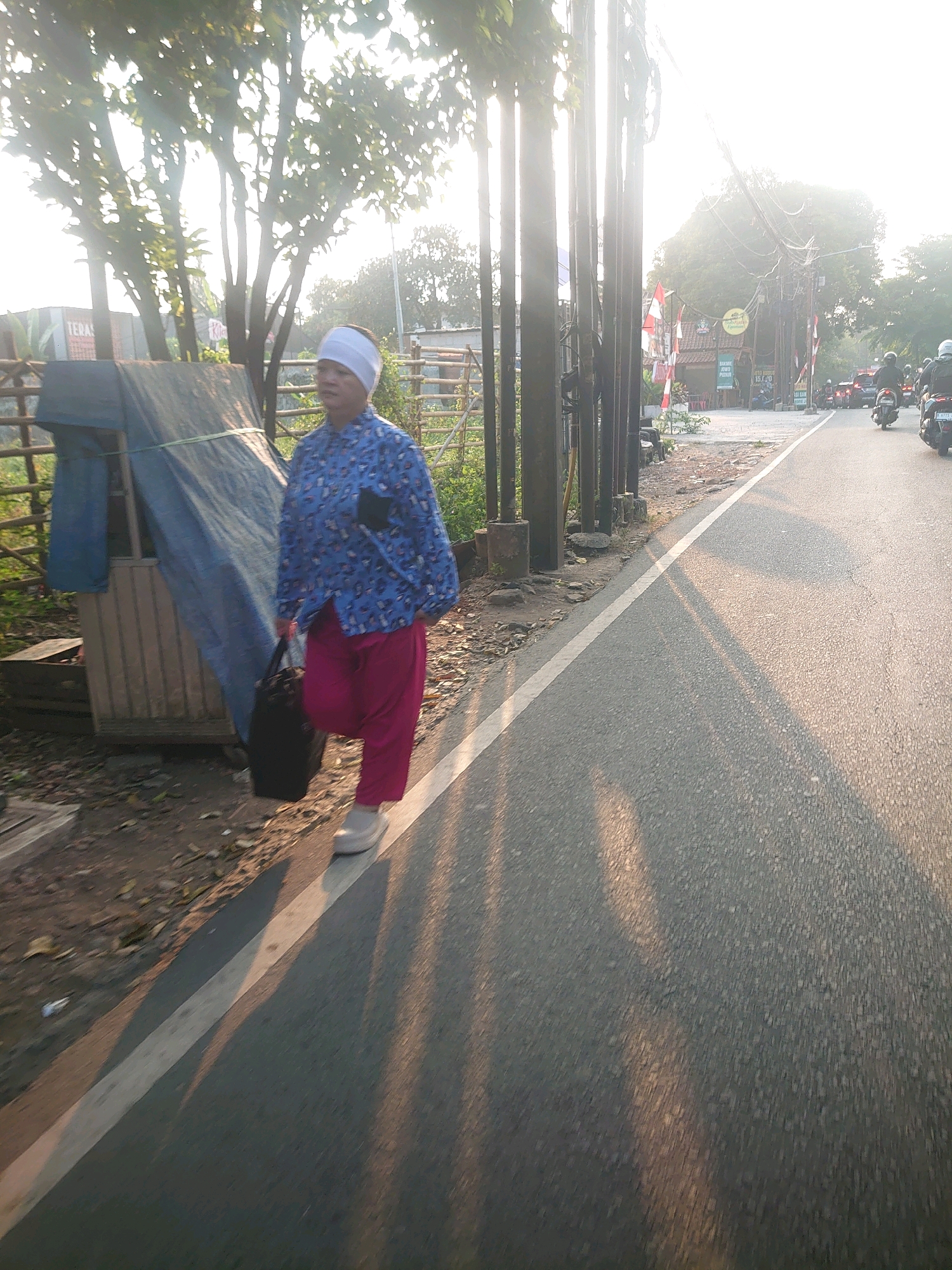 A woman walks along a paved road in South Jakarta, Indonesia, in the late afternoon. She is wearing a blue patterned shirt and bright pink pants, with a white headscarf. She carries a black bag in her left hand. To her left is a small, wooden structure covered with a blue tarp, and beyond that are trees and foliage. The road is lined with utility poles and some traffic is visible in the distance. The sun is low in the sky, casting long shadows across the road.