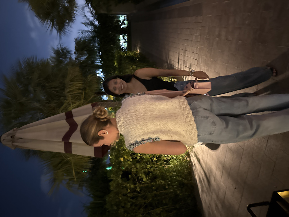 The image displays an outdoor night scene in Dubai, United Arab Emirates. Two women are present on a paved walkway or patio. The woman in the foreground is seen from behind, facing slightly left, wearing a cream-colored knitted sleeveless top with blue trim and light blue jeans. Her hair is styled in a bun. Behind and to her right, a second woman with dark, shoulder-length hair smiles directly at the camera. She is dressed in a black sleeveless top, dark blue jeans, and dark sandals, carrying a light pink shoulder bag with a gold clasp on her right side and a silver-toned watch on her left wrist. Dominating the upper center background is a large, folded patio umbrella, beige with maroon stripes. The environment features lush green hedges and mature palm trees under a dark blue night sky. The ground is made of light-colored rectangular pavers. Artificial light sources illuminate the scene, casting shadows.