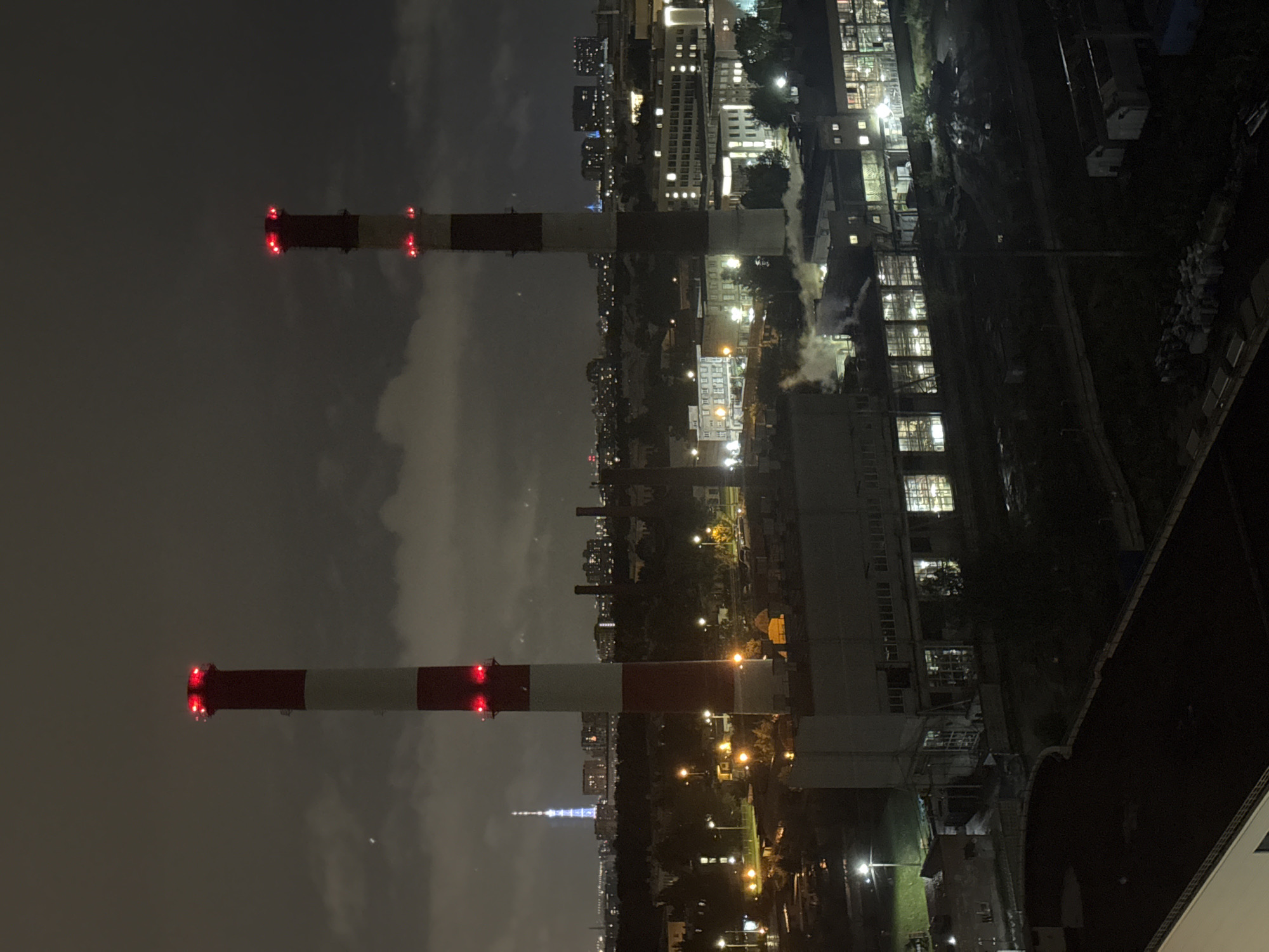 An elevated night-time view shows two industrial smokestacks at the Sinyavino, Russia power plant. The smokestacks are red and white striped with red lights at their tips. The smokestacks are in the foreground, with the city visible in the background. The city is lit up with streetlights and building lights. The sky is dark and cloudy. Steam is coming out of the smokestacks and the buildings. The scene is industrial and urban, with a sense of nighttime activity.