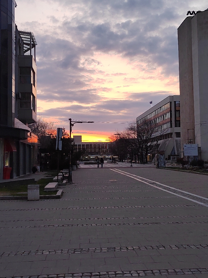 An urban pedestrian street scene in Blagoevgrad, Bulgaria, is captured at sunset. The location is identified as Macedonia Square, looking towards the south-east, with the Municipality building on the right.
The sky exhibits a gradient of colors, transitioning from a vibrant orange and red glow at the horizon, indicative of a sunset, to soft pinks and purplish-grey clouds towards the zenith. Bare, deciduous trees line sections of the street on both sides.
The foreground features a paved pedestrian walkway constructed from rectangular grey and darker grey paver stones arranged in horizontal bands. Two parallel white lines run down the center of this path. On the left, a low concrete planter box with minimal visible greenery and integrated seating is present, alongside a light-colored rectangular trash receptacle.
On the left side of the street, a modern multi-story building with a glass and dark grey facade and prominent balconies is visible. A red awning or architectural element extends from its ground floor. A dark street light pole stands mid-left, adorned with multiple signs.
Further down the pedestrian street, two human figures, appearing as dark silhouettes against the bright sunset, walk away from the viewer. The individual on the left appears slightly taller.
On the right side of the street, a tall, light-colored, multi-story building, identified as the Blagoevgrad Municipality building, stands prominently. It features horizontal bands of windows and a distinctive "M" shaped architectural element on its roof, along with a flagpole flying a blue flag. Below this building, a smaller, angular public sculpture or structure is visible next to a white informational signboard with blue text and a logo.
In the background, a large building with numerous windows, consistent with the American University in Bulgaria (Skaptopara I), is silhouetted against the intense orange and red sunset.