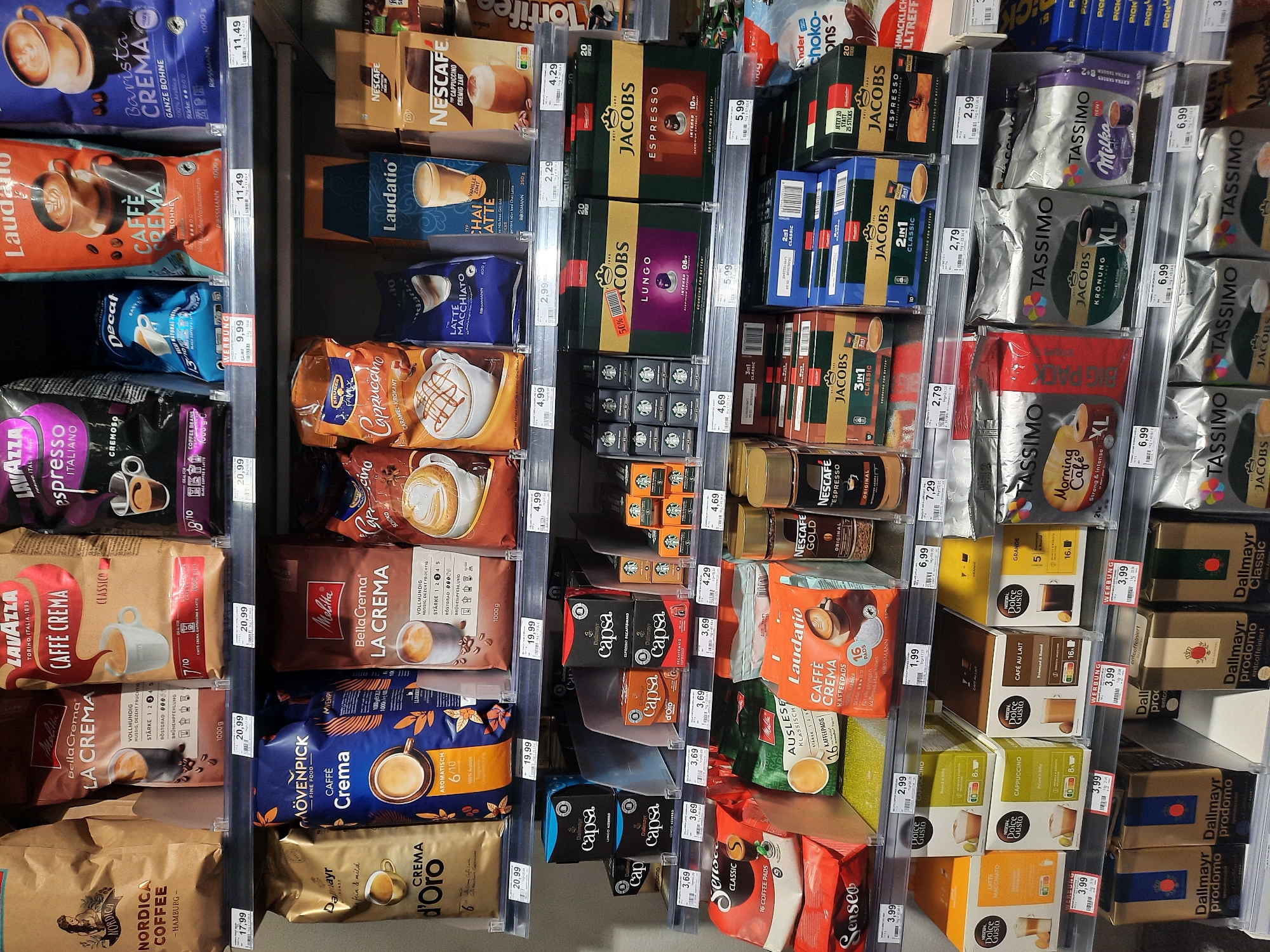 The image displays a shelf in a German supermarket stocked with various coffee products. The brands visible include Lavazza, Melitta, Mövenpick, Jacobs, Nescafé, Dallmayr, Senseo, and Tassimo, among others. The products are mostly packaged in bags and boxes, featuring coffee beans, ground coffee, and coffee pods for machines like Senseo and Tassimo. Prices are displayed on shelf labels in Euros. The arrangement suggests a retail environment with multiple shelves filled with a wide selection of coffee. The location is identified as Rosenheim, Germany, implying a specific retail outlet within that city. There are no people or discernible actions in the image. The environment appears to be a well-lit indoor retail space.