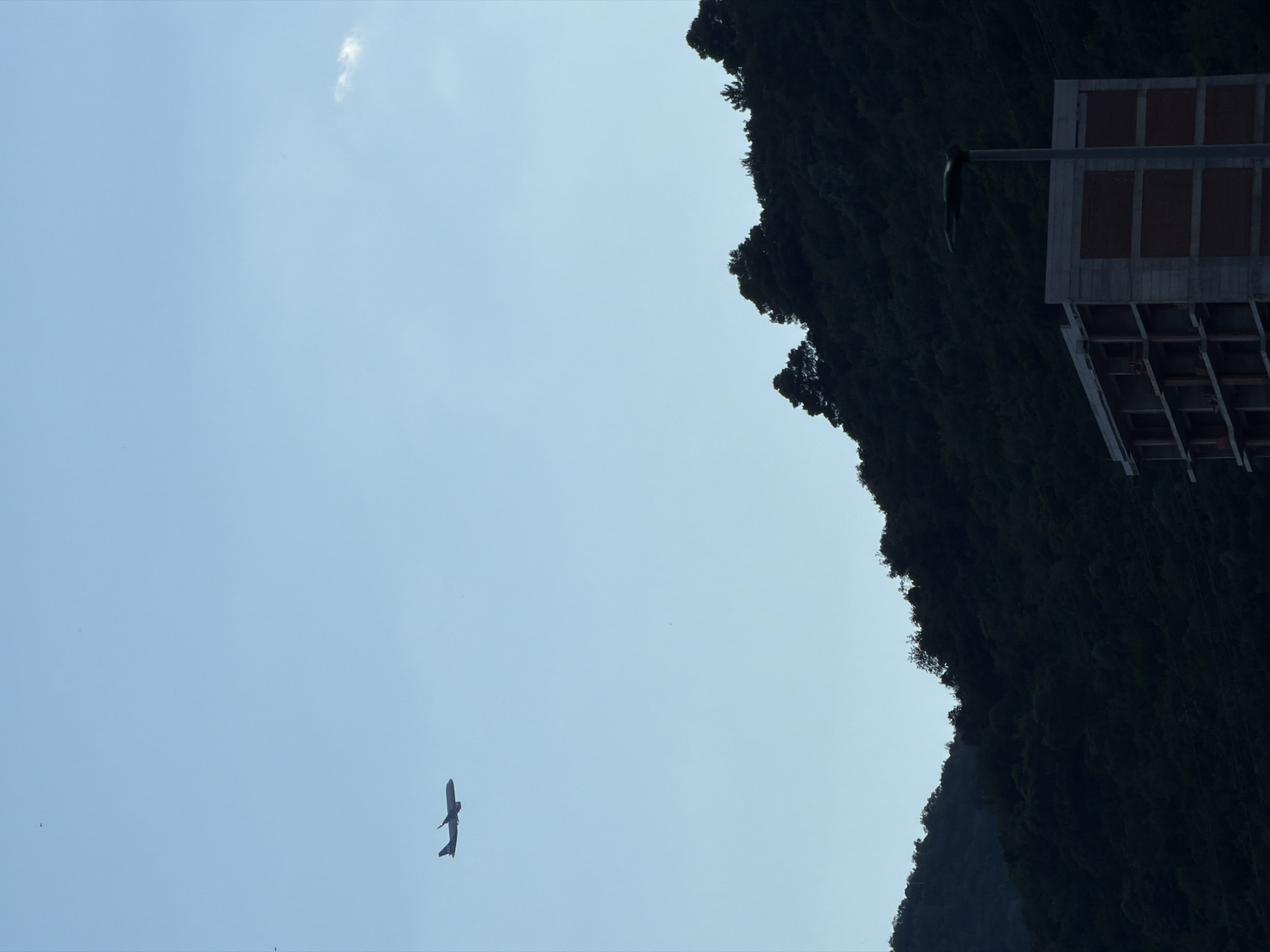 An airplane is flying in the clear blue sky above a dark, tree-covered hillside. In the foreground, there is an unfinished concrete building with bricked-up window openings and a street lamp in front of it. The scene appears to be in Budva, Montenegro.