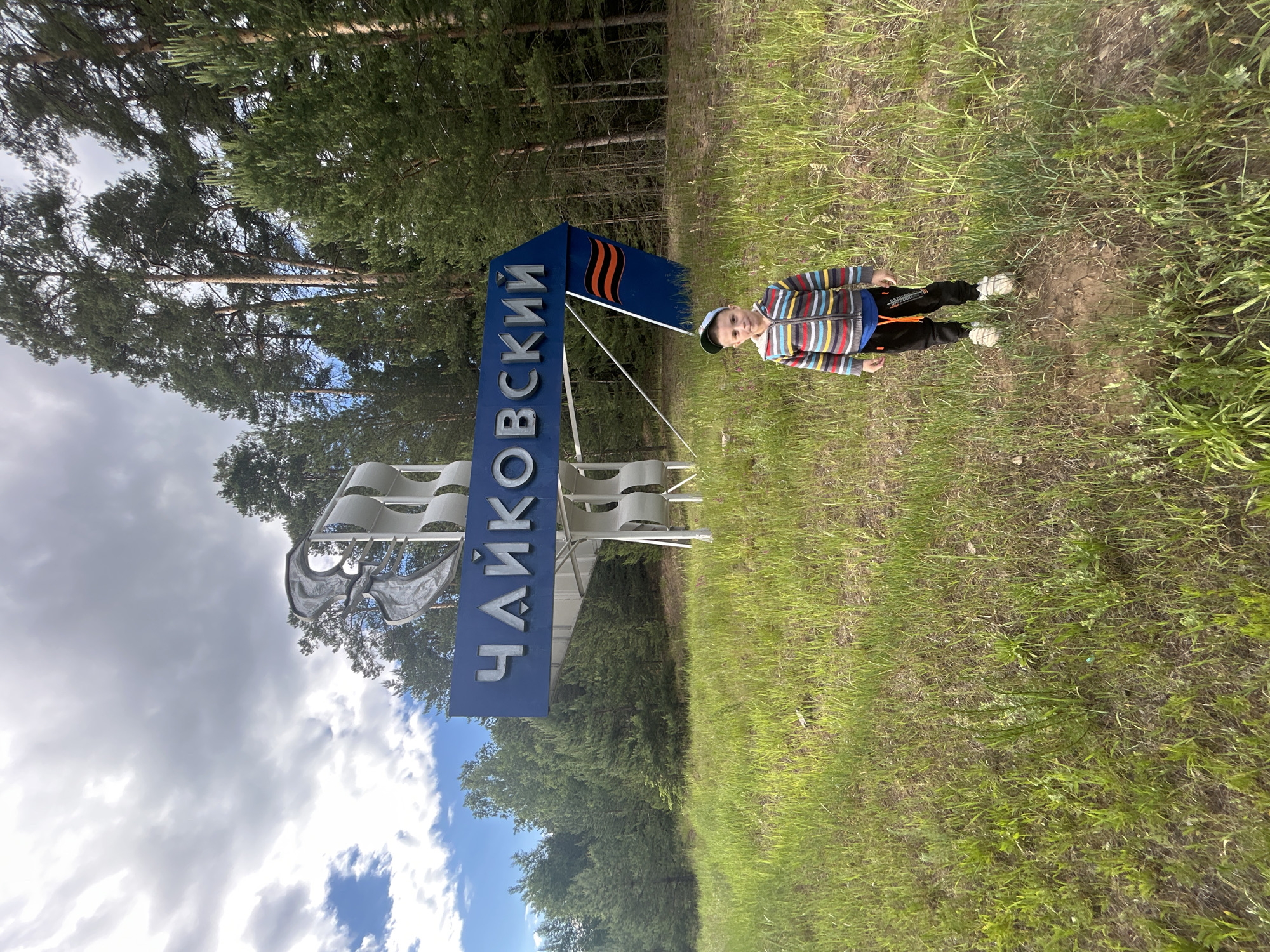 A young boy stands in a grassy field in front of a large sign that says "ЧАЙКОВСКИЙ" in Russian Cyrillic letters. The sign is blue with white lettering and has a decorative metal structure behind it. To the right of the sign, there is a flag with orange and black stripes. The boy is wearing a striped jacket, black pants, and a baseball cap. He is looking at the camera. In the background, there are pine trees. The sky is cloudy with some patches of blue. The scene appears to be an entrance to the city of Chaikovsky, Russia.