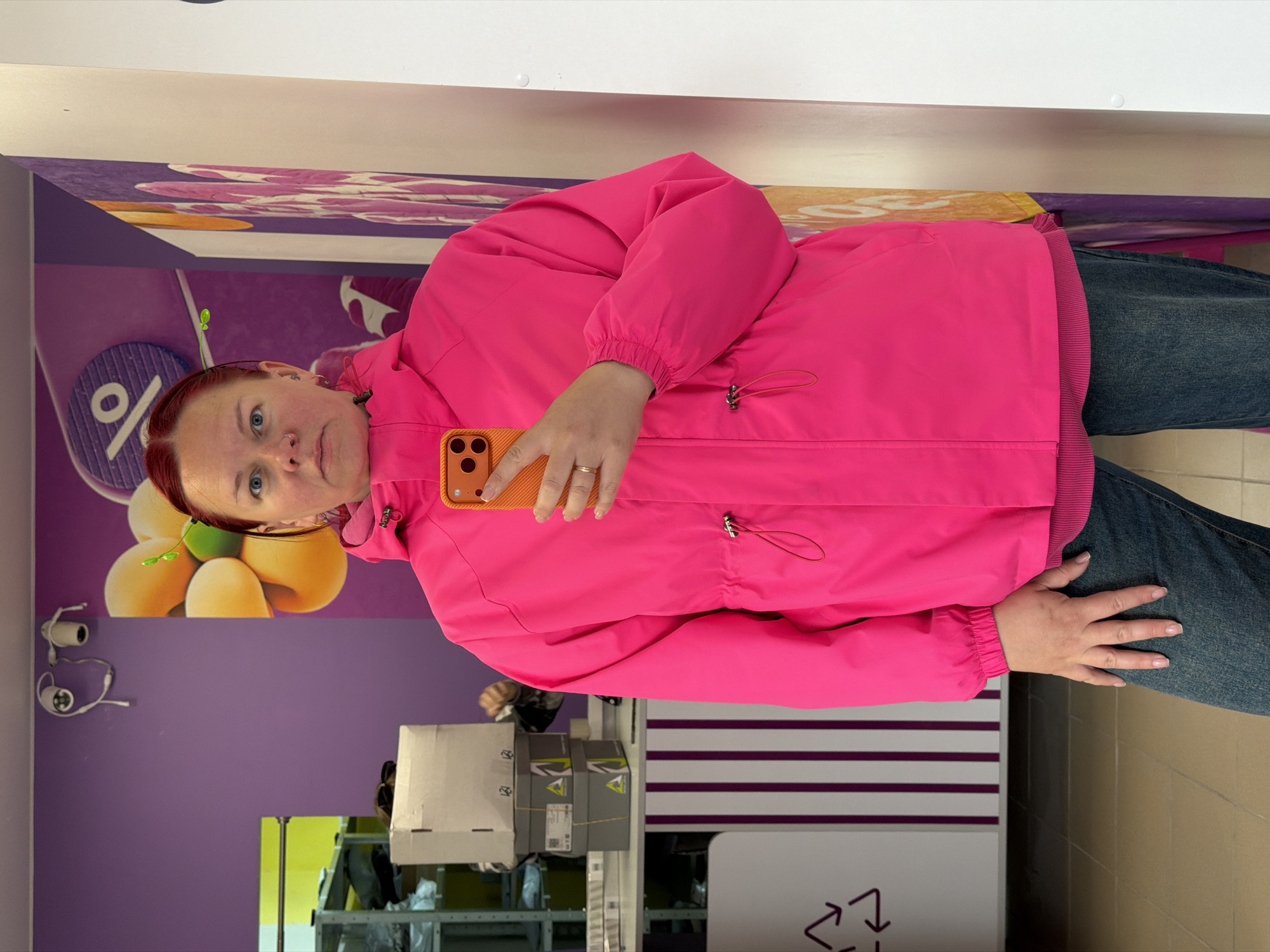A woman with red hair is standing in a store, possibly in the city of Chaikovsky, Russia. She is wearing a bright pink jacket and blue jeans and holding an orange smartphone. She is facing the camera.
The background includes a purple wall with a marketing display featuring fruits, a percentage sign, and other graphic elements. There are shelves with merchandise, possibly in boxes, visible in the lower portion of the image. A recycling symbol is visible on a white bin. The floor has a tiled pattern.