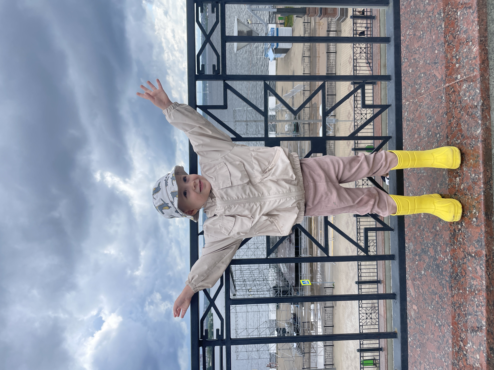 The image shows a young child standing on a stone ledge with arms outstretched against a backdrop of a cloudy sky and a body of water. The child is wearing a light-colored jacket, pink pants, a patterned bucket hat, and bright yellow rubber boots. A black metal railing is in front of the child, and beyond the railing, there is a waterfront area with some structures and a body of water. The railing has a decorative geometric pattern. In the background, there is a large, somewhat complex structure made of scaffolding or metal frames, possibly a stage or temporary seating area, situated near the water. The sky is filled with dark, heavy clouds, suggesting overcast or rainy weather. The foreground consists of a textured, reddish-brown stone surface.
This scene is likely captured on the Izhevsk Kremlin embankment, a popular public space in Izhevsk, Russia, known for its views of the Izhevsk Pond and often used for events and gatherings. The presence of event structures is common in this area during festival seasons.