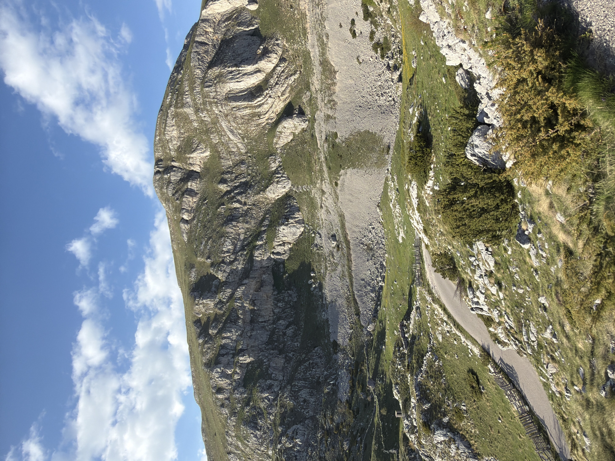 The image shows a mountain landscape in Boričje, Montenegro. A rocky mountain slopes upwards, partially covered with vegetation and a scree slope. To the right, a winding road is visible, edged by a wooden fence in some sections. The sky is blue with scattered clouds.