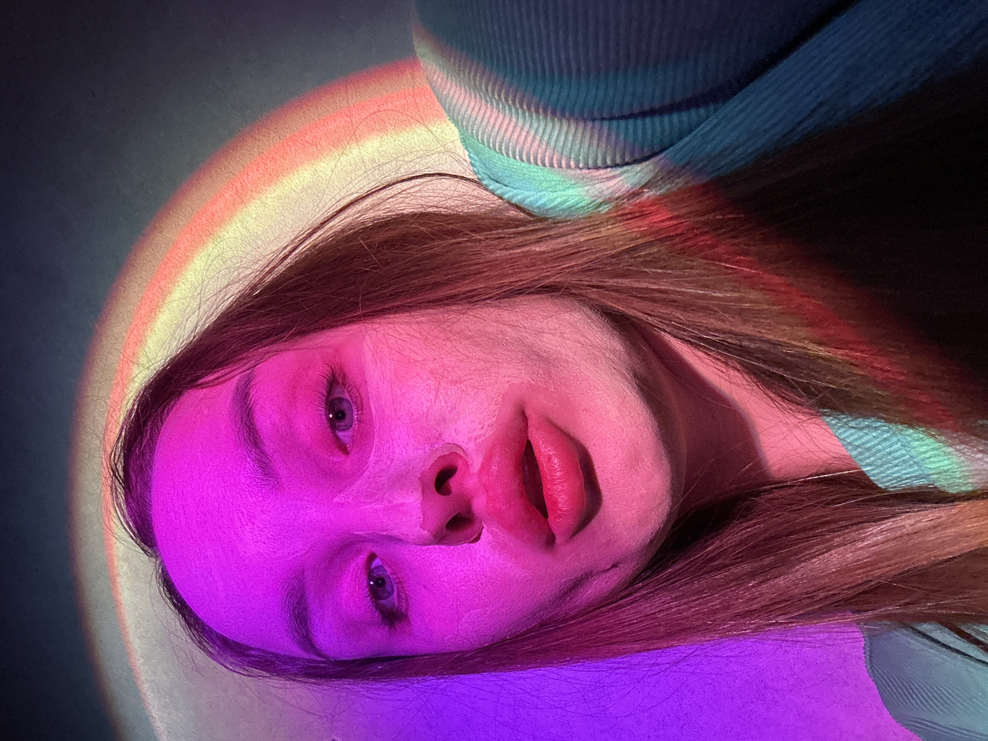 This is a close-up, diagonal portrait of a person, likely female, captured indoors in Chaikovsky, Russia. The subject's head is tilted, with their face illuminated by strong, artificial colorful lights.
The person has long brown hair, parts of which are visible and catch the ambient light. Their face is the central focus, predominantly bathed in vibrant pink and purple light. Their eyes are open and directed towards the viewer. The lips are parted, showing a reddish-pink hue. The skin appears smooth under the intense illumination.
On the left side of the frame, a distinct arc of rainbow-colored light is projected onto a dark background, transitioning from yellow and orange to red. This light also falls partially onto the subject's hair. In the upper right corner, a segment of a blue and turquoise ribbed or textured garment, possibly a sweater, is visible, adding another color element to the scene.
The artificial lighting creates an artistic and moody atmosphere. No specific activity beyond posing for the photograph is depicted. There is no visible text in the image. The time of day and weather are not discernible due to the indoor setting and artificial lighting.
