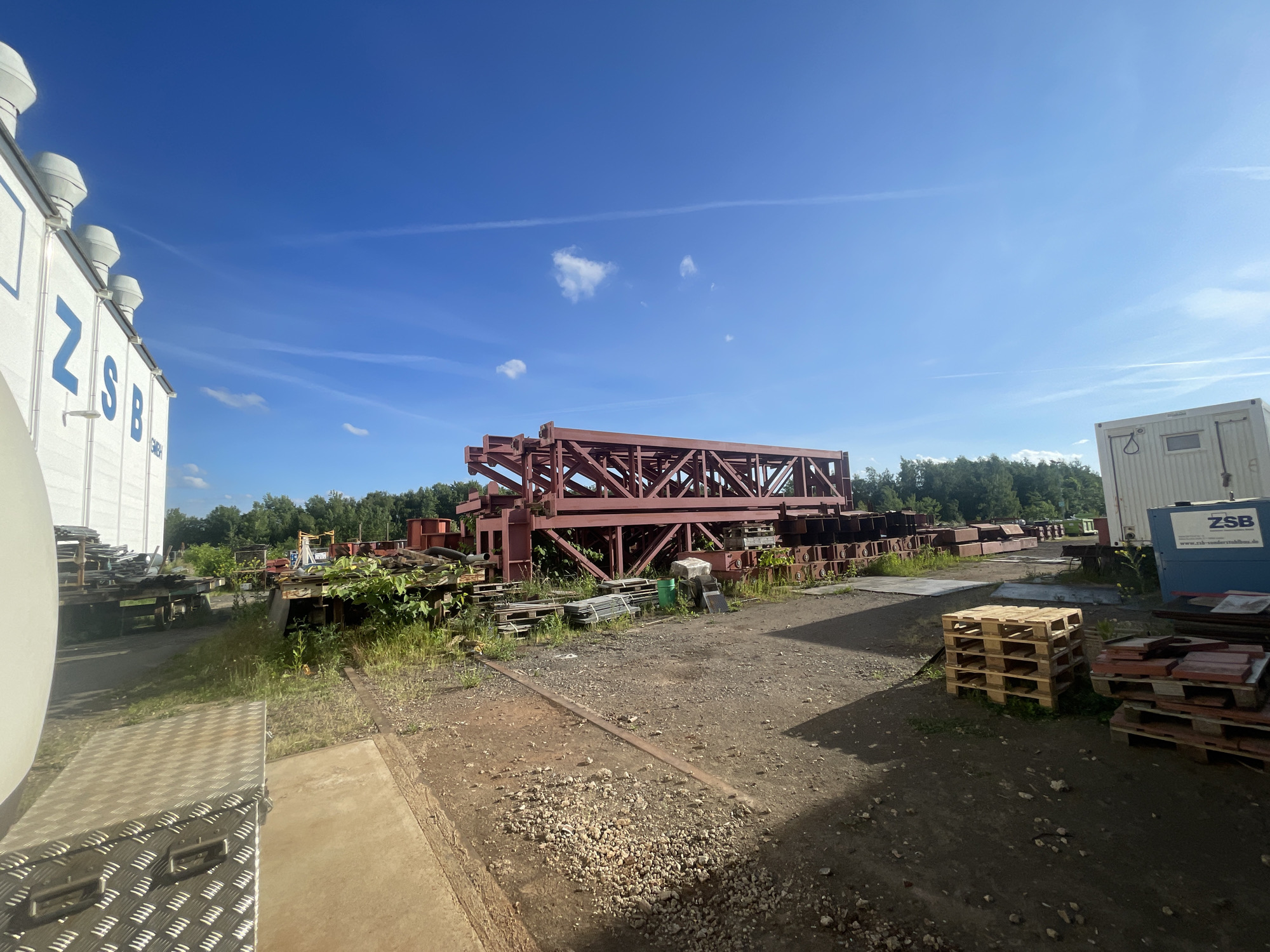 The image shows a storage or industrial yard in the city of Unknown, Unknown, with several large steel structures stacked on the ground. A tall, red-painted steel truss bridge component dominates the center of the image. Around it, there are various metal pieces and pallets. The ground is a mix of dirt, gravel, and concrete pads. In the background, there is a line of green trees. To the left, there's a white building with "ZSB" printed in blue letters. To the right, there is a white shipping container. The sky is blue with a few white clouds.