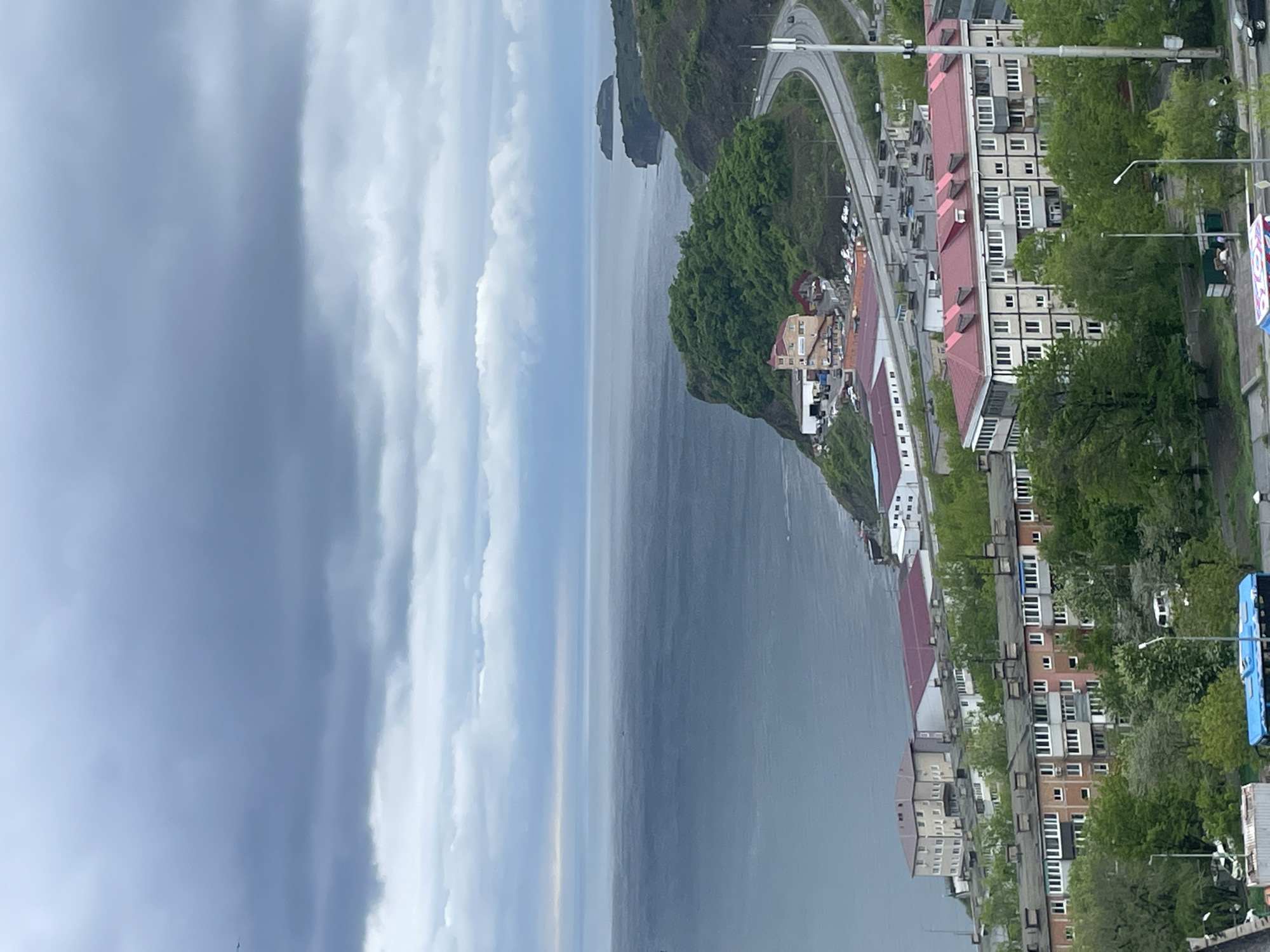 The image depicts a cityscape of Vladivostok, Russia, overlooking a body of water, likely the Sea of Japan. A dramatic sky with heavy clouds occupies the left side of the frame. The right side showcases a coastal road curving around a cliff covered in lush green vegetation. Residential buildings with red roofs and white facades line the street, interspersed with trees. A small island is visible in the distance. The overall atmosphere suggests a rainy or overcast day.