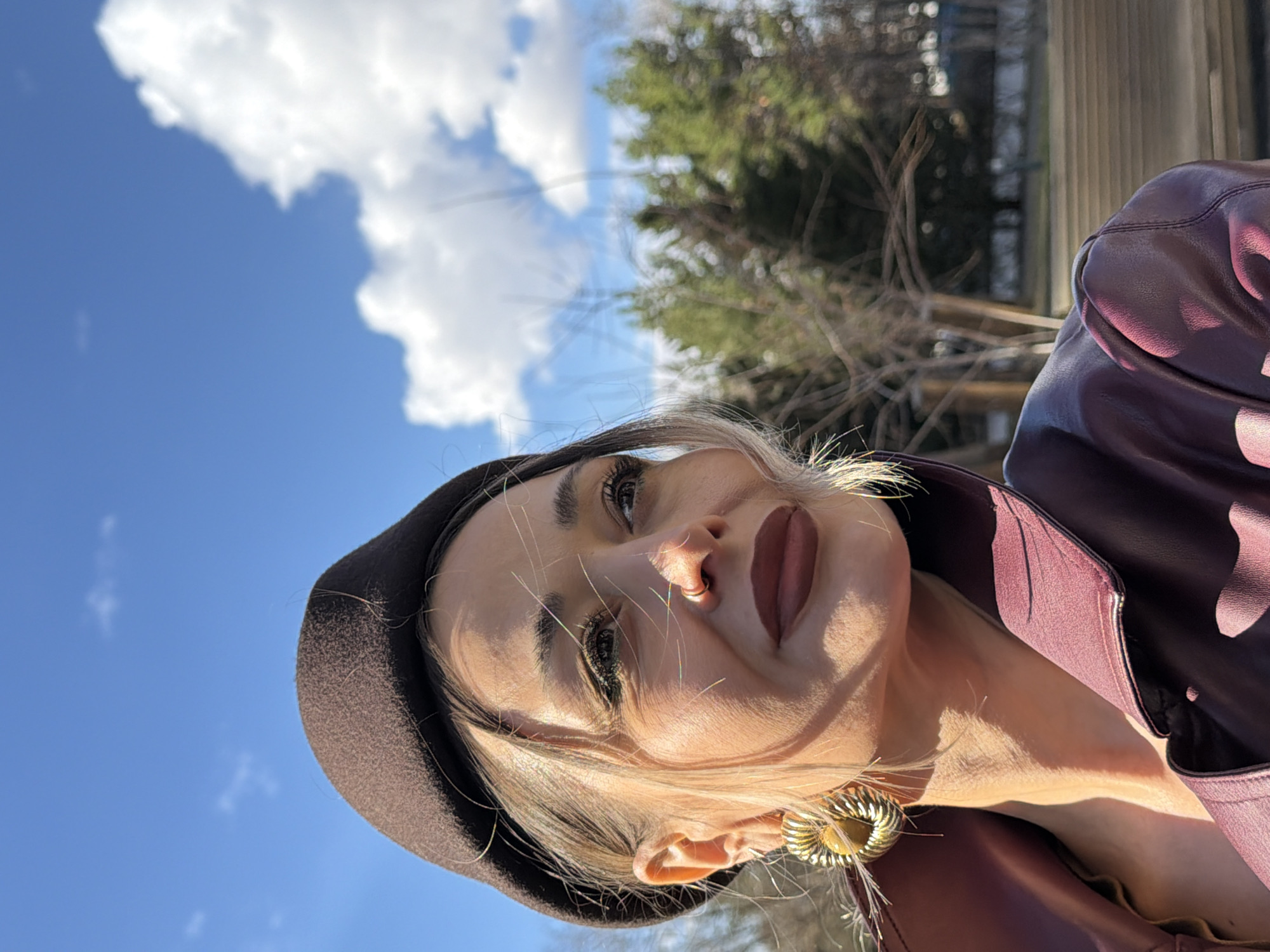 A close-up, upward-angled portrait features a young woman posing outdoors under a clear blue sky in Krasnoyarsk, Russia. She has light-colored hair, dark eyebrows, and is looking upwards with a soft, slightly contemplative expression. Her lips are adorned with a dark, matte lipstick, and a subtle nose ring is visible.
She is wearing a dark brown beret and a deep maroon or burgundy leather-look jacket with a structured collar. A large, ornate golden earring, featuring a ribbed or textured circular design, dangles from her visible ear. Strong, direct sunlight illuminates her face from the left, creating distinct shadows that highlight her features and add depth to the image.
The background consists of a bright blue sky dotted with fluffy white clouds. In the soft-focus distance, bare tree branches intermingle with the green foliage of evergreen trees. A building with classical-style columns can also be vaguely discerned, suggesting an urban or park setting. The image captures a bright, sunny day, emphasizing the contrast between the woman's attire and the natural light. No text is visible.