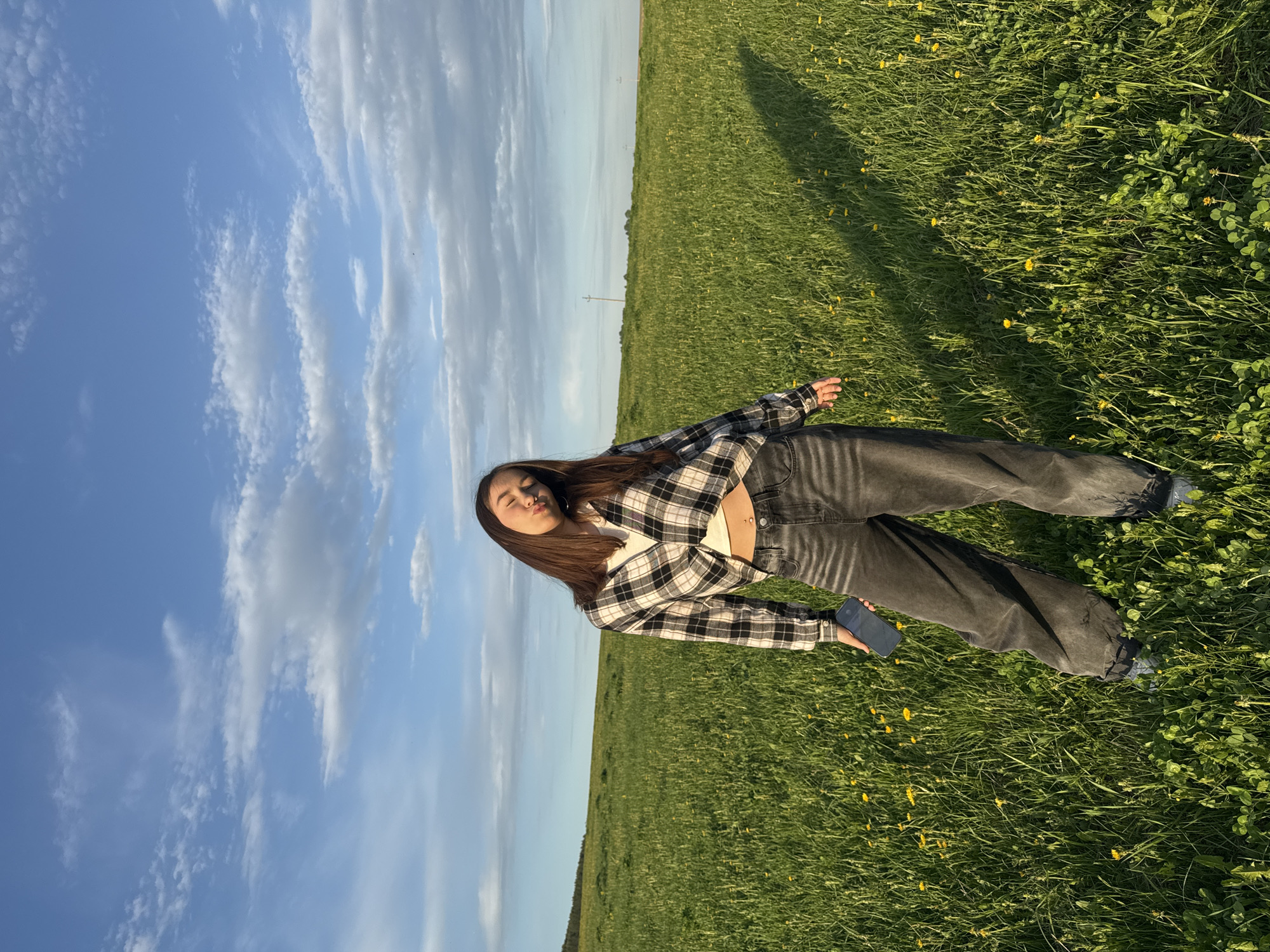 A medium shot shows a young woman standing in a field in Июльское, Russia. She faces the camera at an angle, with her upper body tilted slightly toward the sky. She is wearing a black and white checkered flannel shirt over a white top and dark gray jeans. She holds a dark blue phone in her right hand. Her long brown hair falls down her back and over her shoulders. She has her eyes closed and her lips pursed. The field is green with grass and small yellow flowers. The sky is blue with streaky white clouds.