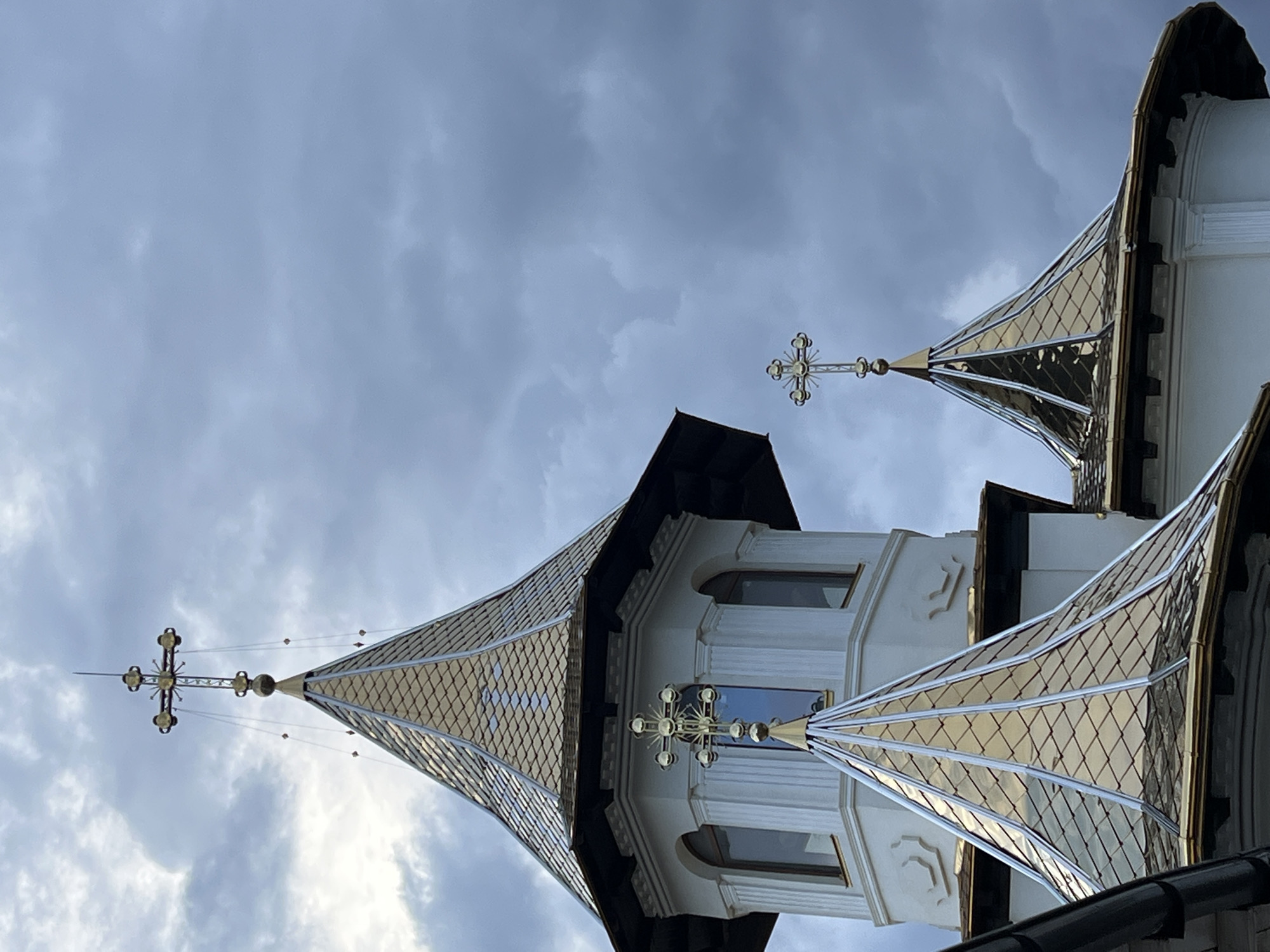 The image shows the gilded spires and crosses of a church or monastery, set against a dramatic, cloudy sky. The architecture features white walls with ornate detailing, arched windows, and pointed roofs covered in gold-colored tiles. Three prominent crosses are visible, each intricately designed with circular embellishments at the ends of the arms. The sky is filled with heavy, dark clouds, suggesting an approaching storm or overcast weather. The perspective is looking upwards towards the structures. The location is identified as Năvodari, Romania. No people or specific actions are depicted in the image.