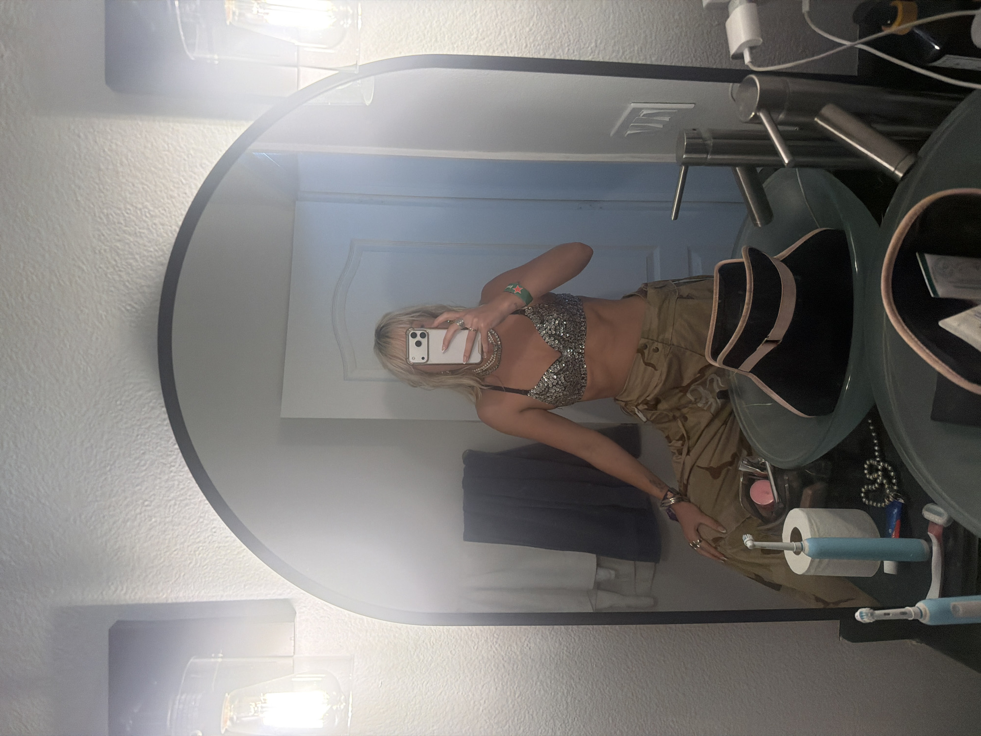 A reflection in a bathroom mirror shows a blonde woman holding a smartphone. The woman is wearing a silver sequined bra top, khaki pants, and jewelry. The bathroom has a white door, a gray towel, a countertop with a sink, and various toiletries including toothbrushes and toilet paper. The location is Palm Desert, USA.
