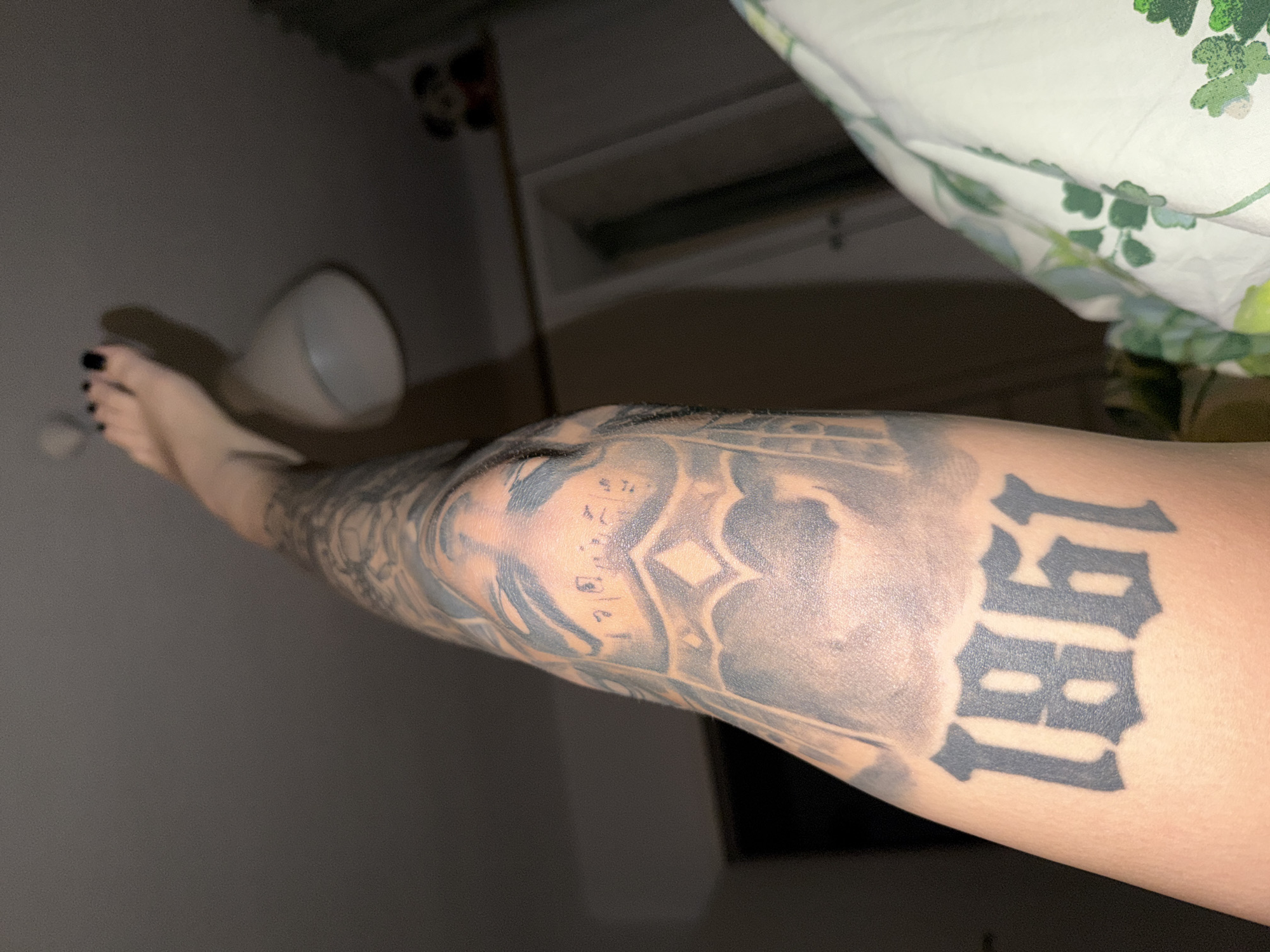 The image shows a close-up view of a person's arm with a tattoo of the numbers "1861" in an old-fashioned font. The arm is tattooed with other imagery as well, appearing to be a sleeve tattoo. The person's feet are visible in the upper part of the frame, with black nail polish on their toes. The background is dimly lit and appears to be an indoor setting, possibly a bedroom, with a white duvet cover featuring a green floral pattern visible on the right side of the frame. A ceiling lamp is visible in the upper right quadrant, and a small Mickey Mouse figurine is perched on a shelf in the background. The scene does not provide any information that can be used to determine the location in the city of Plovdiv, Bulgaria.