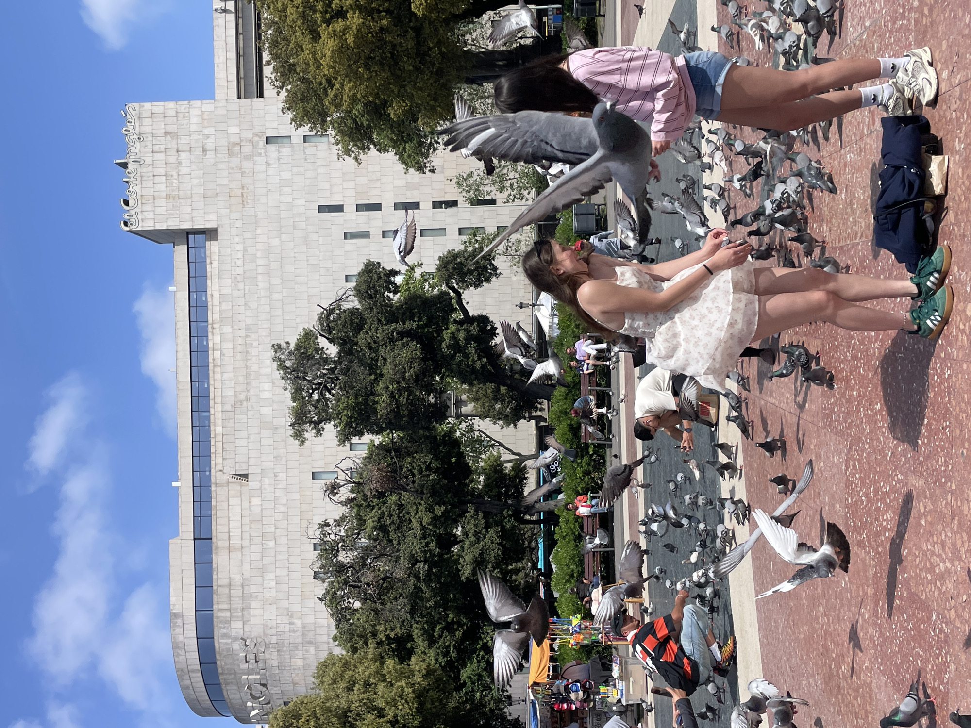 The image depicts a scene in Barcelona, Spain, with a large grey stone building in the background. The building has a rounded corner, with windows on its side. A sign is visible on the corner of the building. In the foreground, two young women are surrounded by numerous pigeons. One woman, wearing a light-colored dress, has her arms outstretched, seemingly interacting with the birds. The other woman is dressed in shorts and a striped shirt. People, benches, and greenery are visible in the midground, suggesting a public square or park. Additionally, some stalls are visible in the scene. The ground is red and textured, possibly made of brick. The sky is blue with a few clouds.