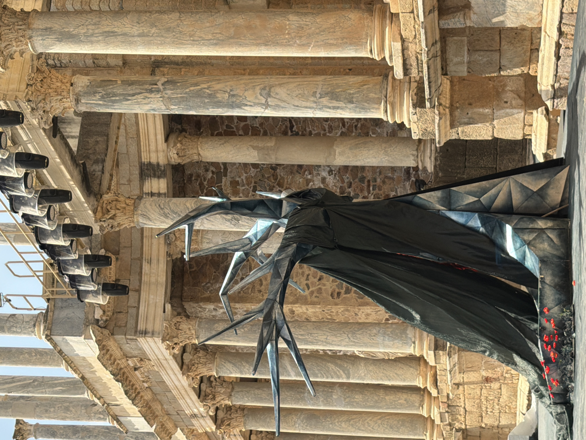 The image shows a theatrical stage set in front of the Roman Theatre of Merida, Spain. The stage features a large, abstract sculpture resembling a skeletal tree with dark fabric draped around its base. Red flowers are planted at the foot of the sculpture. In the background, the stone architecture of the Roman Theatre, characterized by its columns and tiered seating, is visible. Lighting equipment is suspended above the stage area. The scene appears to be set for an outdoor performance.
