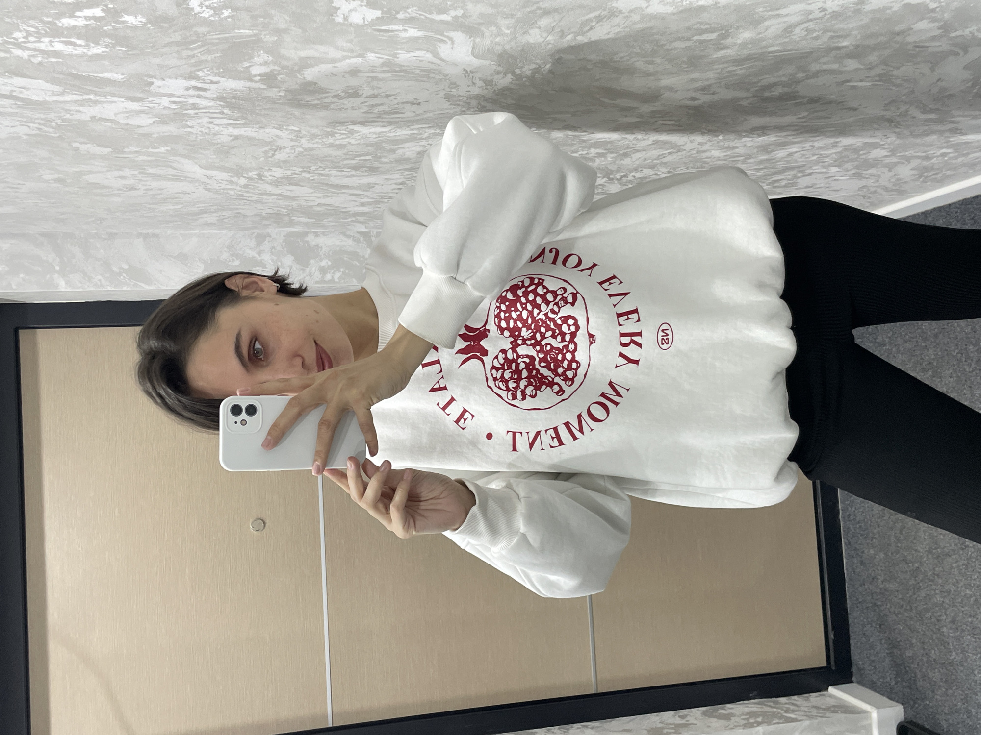 A woman takes a selfie in a mirror. The mirror is framed in black and reflects a tan door with a small round knob. The wall behind the woman is textured and painted white. She wears a white sweatshirt with a circular red design resembling a pomegranate and lettering, paired with black leggings. She holds a white smartphone. Location details are not discernible.