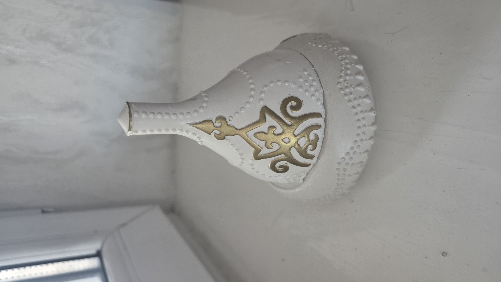A white ceramic decorative item with gold accents rests on a white surface. The item is roughly conical in shape, with a textured base and a smooth, tapered top. The gold design on the body of the item resembles an ornate crest. The background shows a window on the left and a textured wall on the right. The lighting suggests an indoor setting. The image does not provide any information about the city of Chuy, Kyrgyzstan.
