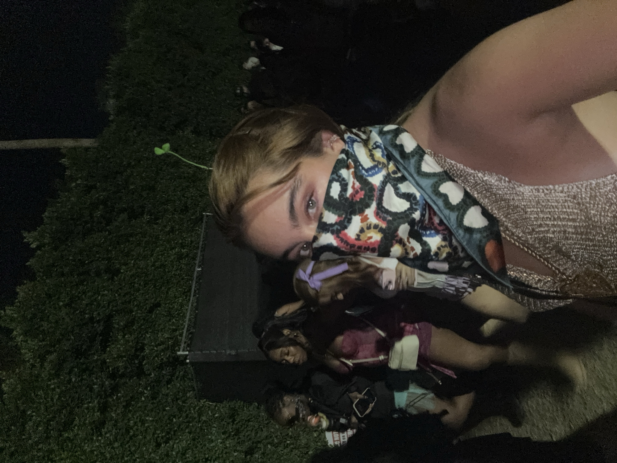 The photograph appears to be taken at night in Indio, United States of America. The focal point is a person partially covering their face with a patterned scarf. They are wearing a shimmering top. In the background, several other individuals are lying on the ground near what seems to be a stage or platform. The environment includes greenery on the left side of the frame.