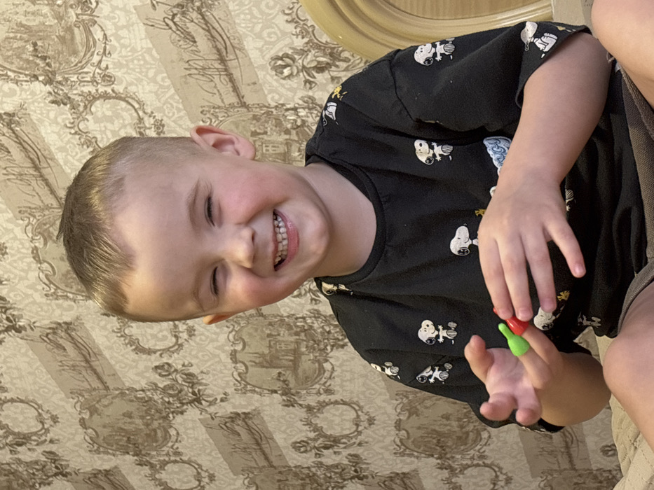 A young male child, approximately three to five years old, is depicted seated indoors in Pavlovsky, Russia. He has short, light-brown hair, fair skin, and rosy cheeks. The child is wearing a black, short-sleeved t-shirt patterned with multiple white cartoon characters resembling Snoopy, and dark shorts. He is smiling widely, with his eyes squinted shut and teeth visible. In his left hand, he holds two small, brightly colored plastic objects; one green piece is positioned on his index finger, and a red piece is held between his thumb and index finger. The background consists of light-toned wallpaper featuring a repeating pattern of framed architectural scenes and ornate floral designs. A segment of a light-colored, possibly wooden or upholstered, furniture piece is visible on the right side of the frame. The child appears to be sitting on a light-colored, soft surface.