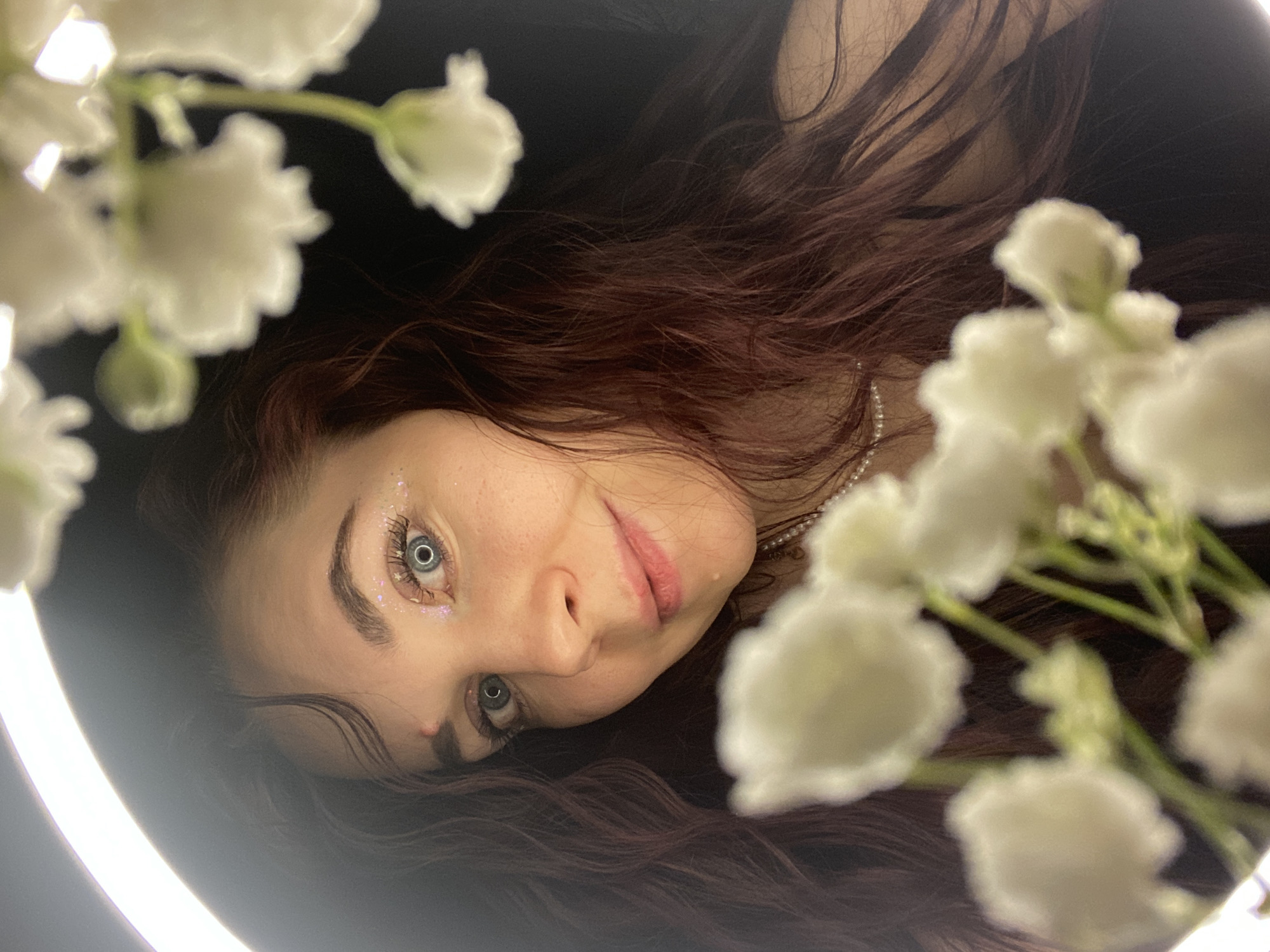 The image presents a close-up portrait of a young woman with long, dark reddish-brown hair, positioned as if lying down and looking towards the camera. Her light blue-grey eyes, enhanced with prominent eyelashes and sparkling glitter makeup around them, are open wide. She has a soft, contemplative expression with slightly parted pink lips. A pearl necklace is visible around her neck/shoulder on the right side of the frame.
The woman's face and hair are illuminated by a bright, circular light source, likely a ring light, which is partially visible on the left side of the image. Soft, out-of-focus clusters of small white flowers, resembling baby's breath, frame her head from the top and right, adding a dreamy aesthetic. The background is uniformly dark, suggesting an indoor setting with controlled lighting. There are no specific location cues visible within the image, but the context provided places the scene in Chaikovsky, Russia. There is no visible text in the image. The overall impression is one of a stylized and artistic photograph.