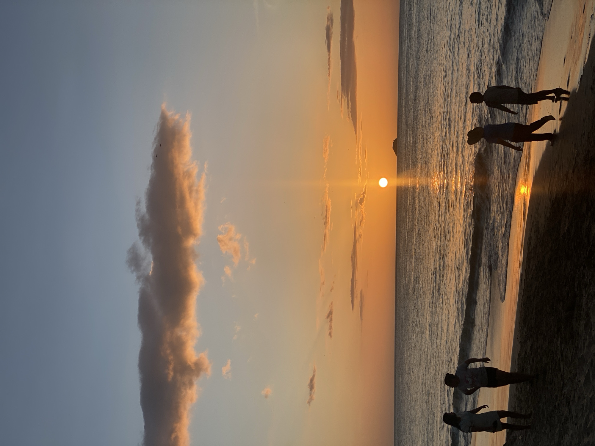 At sunset on a beach in Pedro González, Venezuela, four individuals walk along the shoreline. The sky is a gradient of blue and orange, with clouds tinged with pink and gold. The sun, a bright orb, is low on the horizon, casting a shimmering reflection on the calm ocean waves. In the distance, a small island is visible. The individuals, silhouetted against the vibrant sky, are engaged in leisurely activity on the sand. The scene captures the peaceful end of a day at the coast.