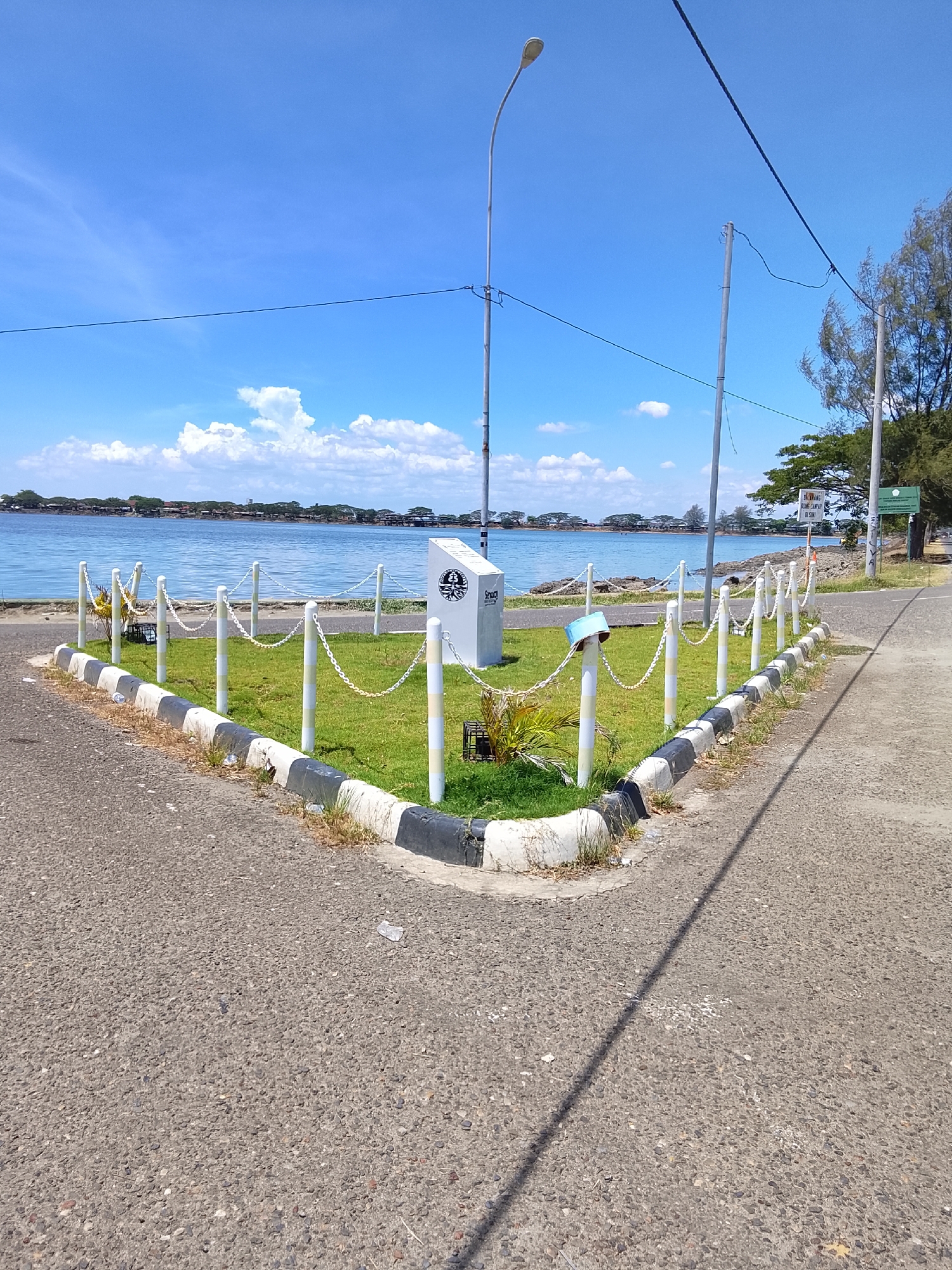 The image depicts a paved area adjacent to a body of water, likely the sea, in Lhokseumawe, Indonesia. In the foreground, a grey asphalt road curves to the right, with a small patch of green grass bordered by a curb made of alternating black and white painted concrete blocks. A white chain-link fence with vertical posts, also painted with alternating yellow and white stripes, encloses a triangular grassy area. Within this fenced area, a white rectangular monument stands, featuring a circular emblem with a stylized design. A street lamp on a tall pole is visible behind the monument, and power lines crisscross the sky. The water, calm and blue, stretches across the mid-ground, with a distant coastline dotted with buildings and trees. The sky is a vibrant blue with scattered white cumulus clouds. The overall scene suggests a public space, possibly a park or promenade, by the water.