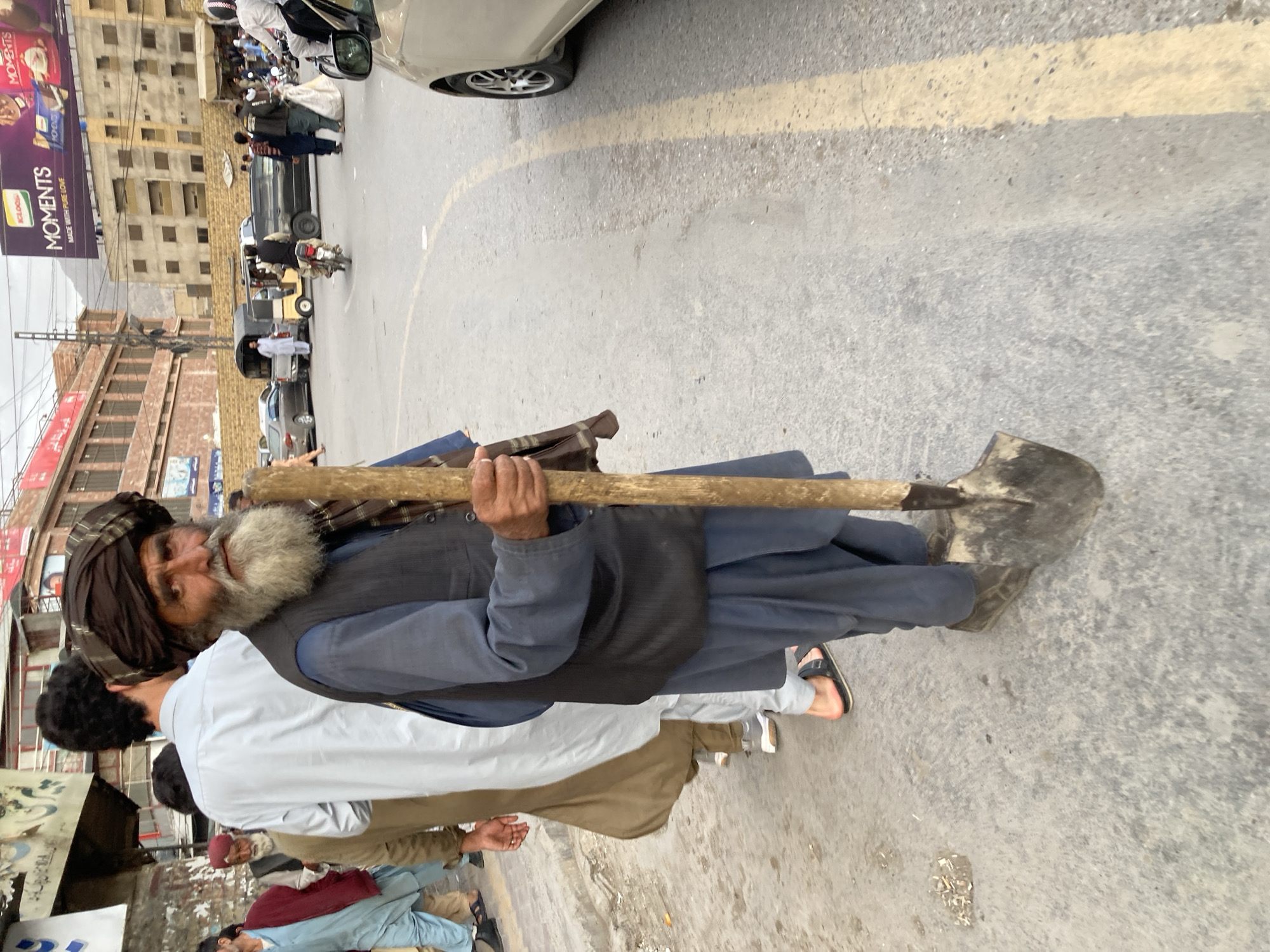 The image captures a bustling street scene in what appears to be an urban area, likely in Quetta City Tehsil, Pakistan, given the context. The photograph is taken from a slightly elevated perspective, looking down and across the street.
The central figure is an older man, positioned prominently in the mid-ground, holding a long-handled shovel. He has a distinguished appearance with a long, thick, grey beard and is wearing a dark brown turban, a dark vest over a blue traditional garment (likely a shalwar kameez), and dark trousers. His hands, with visible wrinkles, firmly grip the shovel's wooden handle. His gaze is directed slightly towards the left of the frame.
In the immediate foreground, partially obscuring the main figure's lower body, is the back of another young man with dark, curly hair, wearing a white shirt and light brown trousers. Other men are visible further back, dressed in traditional attire, some walking or standing.
The street itself is a concrete or asphalt road with a faded yellow line marking. Traffic includes several motorcycles, one with a rider, and an auto-rickshaw. A light-colored car is partially visible on the right edge. Buildings line both sides of the street; those on the left appear to be multi-story commercial or residential structures with numerous windows, some appearing aged.
A large billboard is prominent in the upper left, displaying the brand name "MOMENTS" multiple times, along with "MADE WITH PURE LOVE" and "KELLOGG'S." The sky is overcast, contributing to a muted, diffused lighting throughout the scene, suggesting daytime. The overall impression is a vibrant, everyday street activity typical of a South Asian city.
