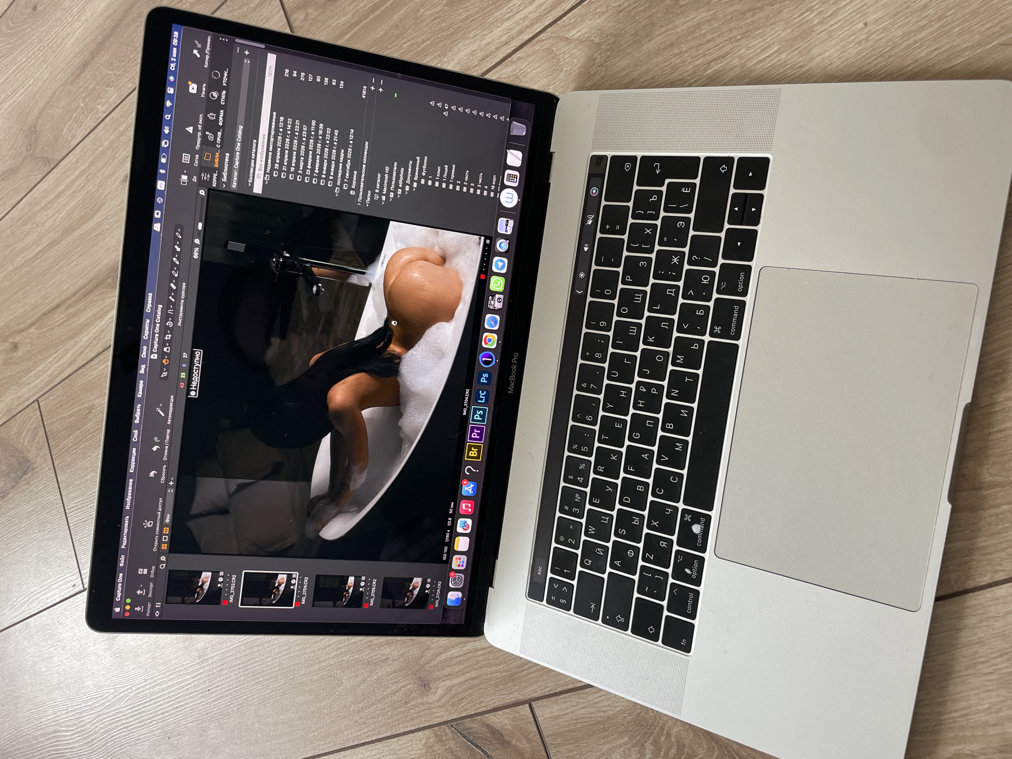 A silver MacBook Pro is open on a light wood floor. The screen displays the image editing software "Capture One", with an image of a person in a bathtub with bubbles loaded in the editor. The image files "IMG_2702.CR2", "IMG_2704.CR2", "IMG_2705.CR2", and "IMG_2706.CR2" are displayed in the bottom toolbar of the application. The right side of the screen shows the file directory and some basic image processing operations.
The keyboard layout appears to be in Russian Cyrillic, with some keys labeled in both English and Russian. The MacBook Pro is a modern model, indicated by the thin bezels around the screen and the large trackpad. The location is the city of Kazan, Russia, as indicated by the Cyrillic letters on the keyboard.