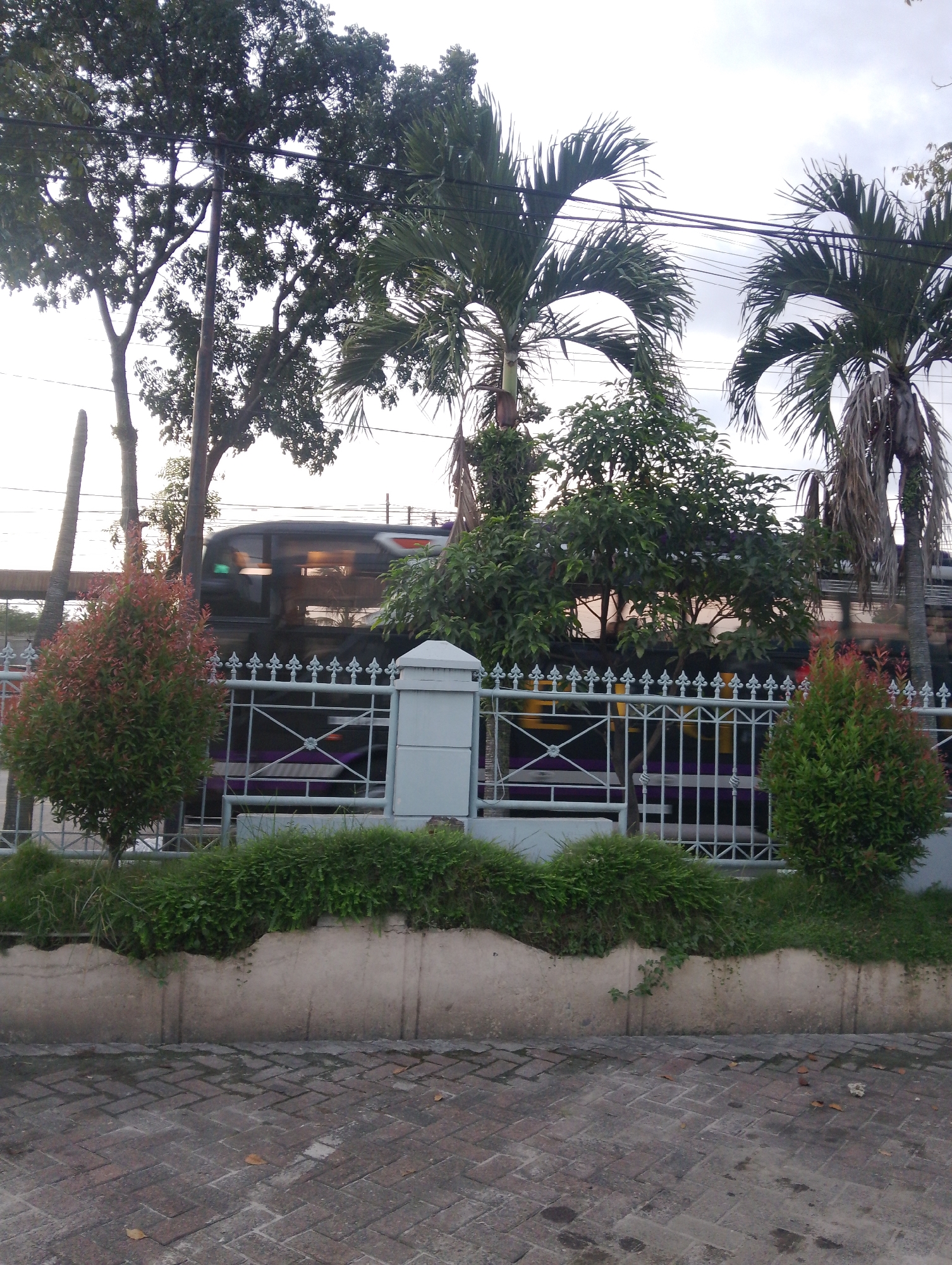 A bus travels past a white metal fence with decorative elements, overgrown green bushes, and palm trees. The bus, dark colored with tinted windows, shows interior lighting and some blurry movement. The fence encloses a walkway with a concrete border and a brick-paved surface. The sky above is overcast. The scene appears to be on a street in Pematang Siantar, Indonesia.