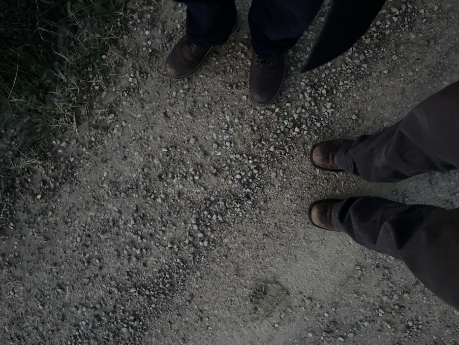 Three individuals are depicted from a high-angle perspective, showing only their lower legs and feet on a gravel path. To the left, one person stands with dark blue trousers and brown lace-up shoes. To the right, two more individuals are positioned; one wears dark grey or brown trousers and sturdy brown boots, while the other's legs and feet are partially visible, also in similar attire.
The scene takes place outdoors on a path composed of small grey and brown gravel mixed with dirt. Sparse patches of green and dried grass line the left edge of the path, suggesting a rural or semi-rural setting. The lighting is dim and diffused, indicating a time of day like twilight or early evening, with soft, ambient light and an absence of harsh shadows. The individuals appear to be standing still, observing their surroundings in Montalcino, Italy. No visible text is present in the image.