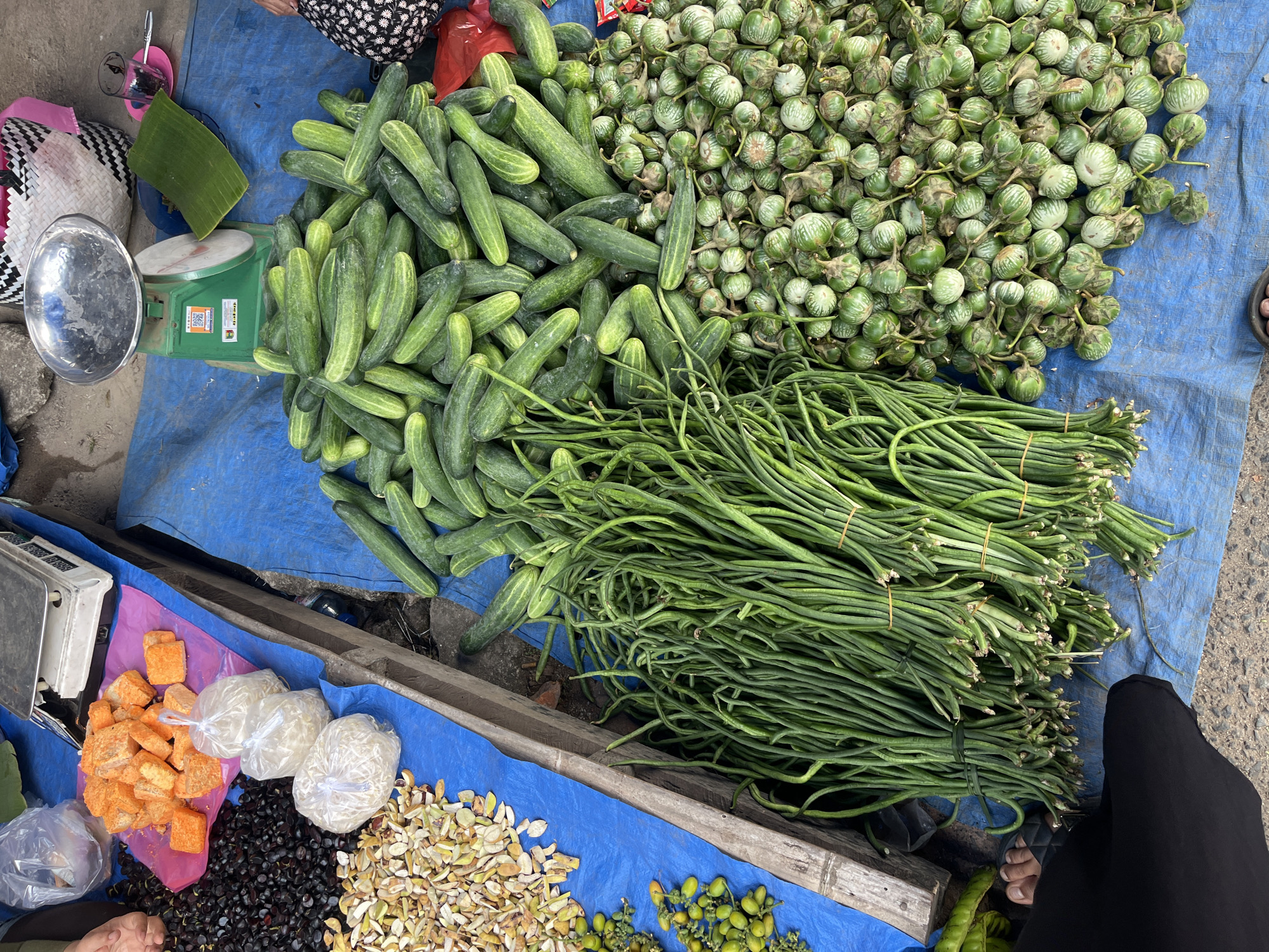 The image depicts an outdoor market scene in Kemuning, Indonesia, likely a street market or a local produce vendor. The foreground is dominated by a blue tarp spread on the ground, serving as a display surface.
On the left side of the blue tarp, a wooden structure, possibly part of a market stall, is visible. Piled on this structure are various items. In the upper left, there are small, orange, cube-shaped items, possibly candied fruit or spices, covered by a clear plastic bag. Below these are dark, round objects, likely berries or small fruits. Further down, there are piles of light-colored, split seeds or nuts. To the left of these seeds, a cluster of small, green, oval fruits is visible, some with yellow tinges. Three white, granular items, possibly bagged spices or grains, are also present on the wooden structure.
The majority of the blue tarp is covered with fresh produce. In the center and right of the tarp, large bunches of long, green beans are tied together with elastic bands. To the right of the beans, a substantial pile of cucumbers is visible. Further to the right, a mound of small, round, green eggplants is displayed.
In the background, a green and silver weighing scale sits on a blue tarp. Behind the scale, a woven basket with a pink interior and a small glass with a pink saucer are present. A single green leaf, possibly a banana leaf, is placed on the tarp near the scale.
Parts of people are visible. In the lower left, the feet of a person wearing dark footwear are visible. In the far right, the patterned fabric of clothing worn by another person is partially visible.
The overall scene is one of a vibrant, bustling market, with a focus on fresh produce. The lighting suggests it is daytime.