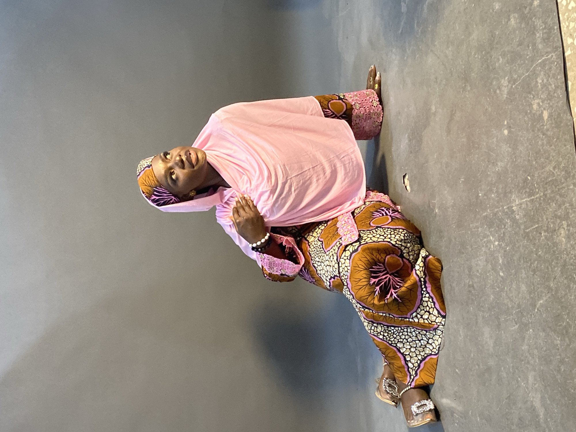 An image, captured indoors in Jos, Nigeria, features a woman posing against a plain dark grey backdrop and floor. The photograph is currently oriented horizontally, but the subject appears to be in a seated or kneeling position, rotated 90 degrees clockwise.
The woman is wearing a light pink head covering, possibly a hijab or large scarf, that drapes over her shoulders and upper body. Her main garment is a vibrant, ankle-length dress or two-piece outfit with an intricate, multi-colored pattern. The dominant colors in the pattern are brown, white, and various shades of pink, forming large, organic, and abstract designs that might resemble stylized flowers or cellular structures. The sleeves of her outfit have a delicate pink lace trim. On her left wrist, she wears a black and white beaded bracelet. Her feet are visible, clad in light-colored shoes or sandals adorned with prominent, sparkling buckles or embellishments.
The woman's complexion is dark. She is looking upwards and slightly to her left (viewer's right in the rotated image), with a gentle, serene expression and a faint smile. Her mouth is slightly open. Her hands are resting in front of her, beneath the pink head covering. The background and floor are uniformly dark grey, providing a stark contrast to her colorful attire. There is no visible text in the image.