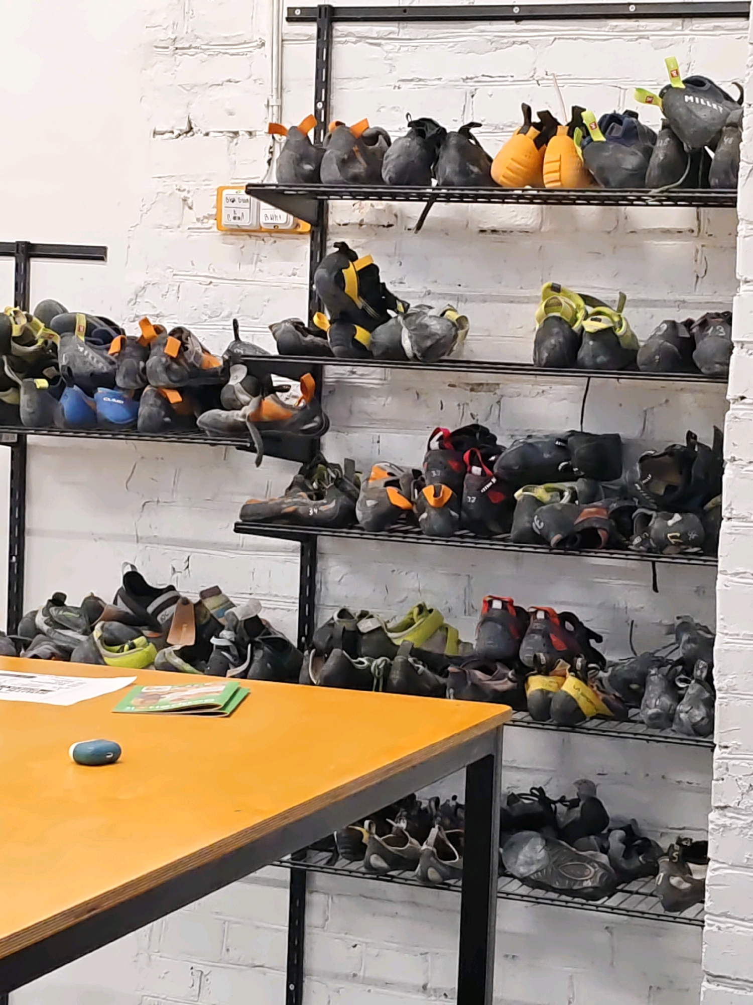 The image depicts a shelving unit filled with climbing shoes. The shoes are varied in color and style, and are organized on multiple shelves. Some shoes are displayed in pairs, while others are single. The shelves are made of metal and are mounted on a white brick wall. A wooden table with some papers and a small object is visible in the foreground. The scene appears to be an indoor climbing facility. There is no indication of the city of Rostov-on-Don, Russia, in the image.
