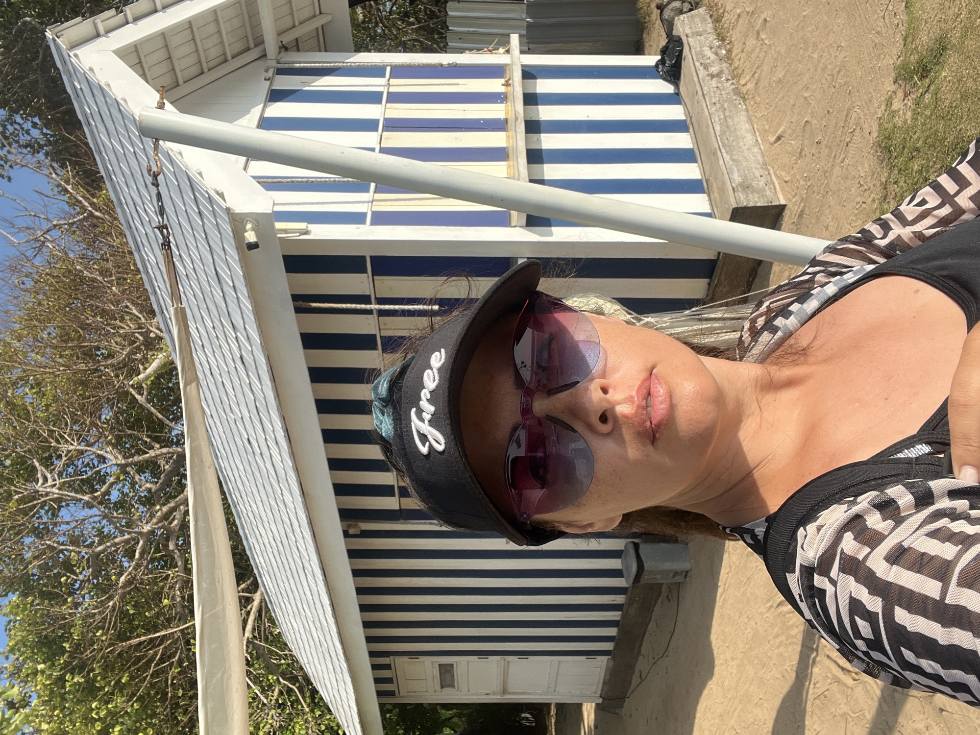 A medium shot captures a woman taking a selfie on a sandy beach in Kuta Selatan, Indonesia. She is wearing sunglasses, a black visor with the word "Free" embroidered in white, and a black and white patterned cover-up over a black tank top. Behind her, a small, striped building with blue and white vertical lines serves as the backdrop. The building has a white-shingled roof and a white door on the left side. Trees with green leaves and bare branches are visible above the building. The ground is covered in sand, with some patches of green grass. The lighting suggests it is daytime, possibly late afternoon, due to the warm glow on the woman's skin.