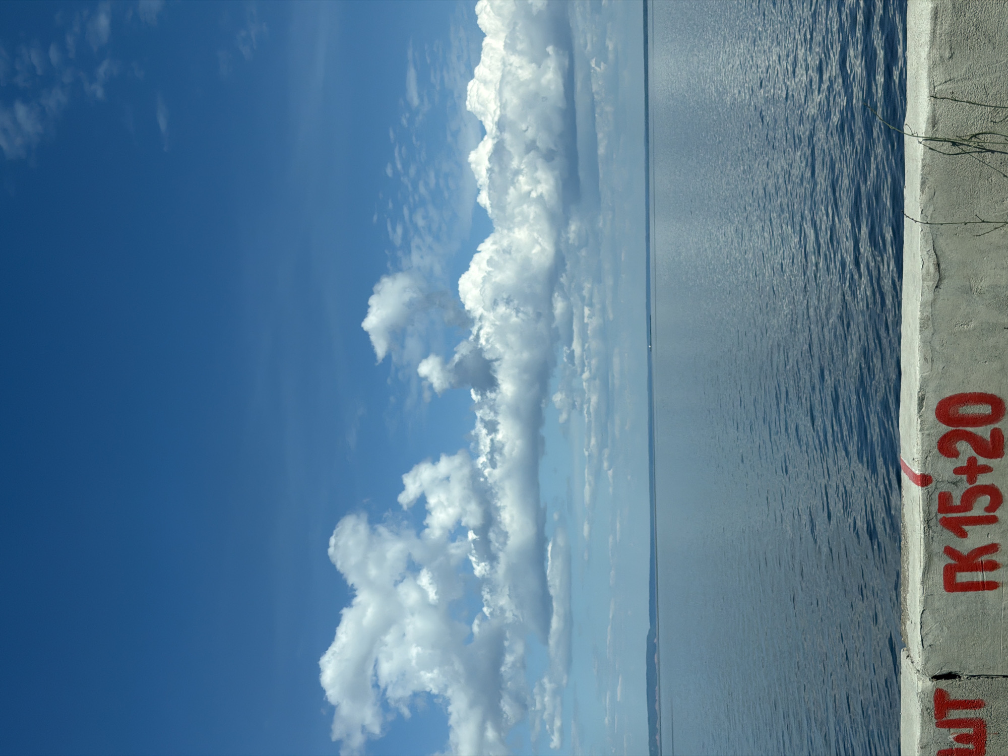 The image displays a panoramic landscape view over a large body of water, located in the city of Chaikovsky, Russia, likely depicting the Kama Reservoir. The upper two-thirds of the frame are dominated by a vibrant blue sky featuring numerous white cumulus clouds, varying in size and density, with some appearing more developed and others as wispy formations. The lower third of the image shows an expansive, calm body of blue-grey water with subtle surface ripples. A faint, low-lying landmass is visible along the distant horizon on the left side. A small, indistinct white object, possibly a boat or buoy, is faintly visible on the water near the far left horizon. In the immediate foreground, a grey concrete barrier or wall extends horizontally across the bottom of the frame. This barrier features red graffiti, including the partially visible Cyrillic characters "ШТ" on the left and "ПК 15+20" prominently in the center. A vertical red line mark is also present on the barrier to the right of the numerical graffiti. Sparse, dry vegetation protrudes from the base of the concrete barrier on the right. The scene is illuminated by bright daylight. No human figures or discernible actions are present.