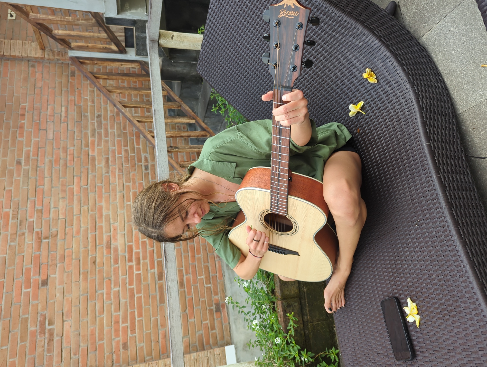 A woman is holding a guitar while seated on a wicker chair in North Kuta, Indonesia. The guitar, branded "Bromo" at the headstock, is oriented vertically in front of the woman. She is wearing an olive-green shirt and dark shorts, and she is barefoot. The surrounding environment includes a brick wall to the left, a wooden staircase in the background, and greenery with white flowers near the bottom of the frame. A black smartphone and two yellow flowers rest on the wicker chair beside her. The flooring is gray concrete.