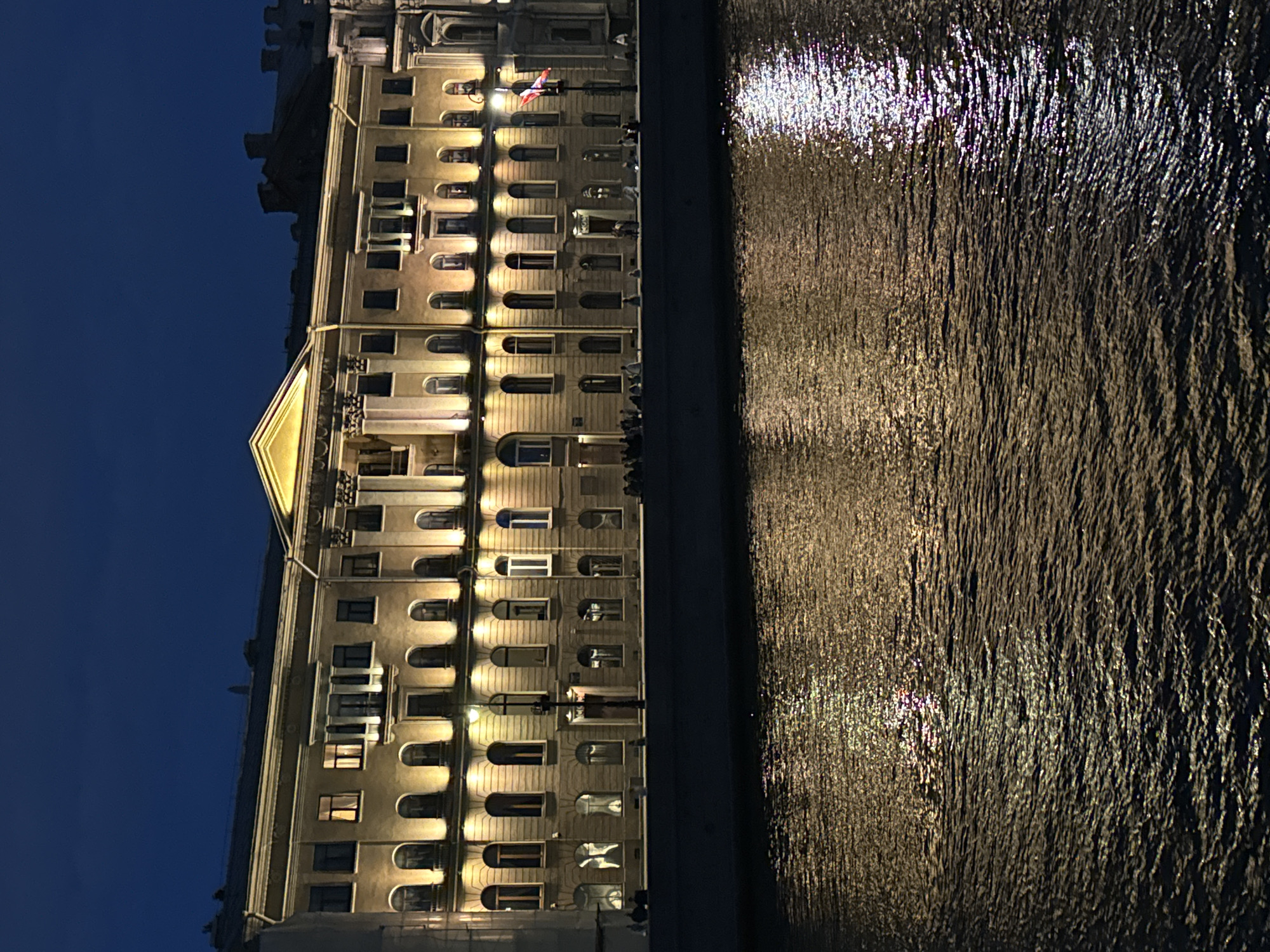 The image shows a large, illuminated building on the embankment of a river at night. The building, which appears to be classical in style with columns and arched windows, is lit from below, casting a warm glow on its facade. Reflections of the building's lights shimmer on the dark, rippling surface of the water. Several people are visible on the embankment, some standing and others seated. A flagpole with a red and white flag is mounted on a pole in front of the building. The sky is a deep, dark blue, indicating it is nighttime. This scene is likely Saint Petersburg, Russia, given the architectural style and the presence of a prominent embankment and river, common features of the city.