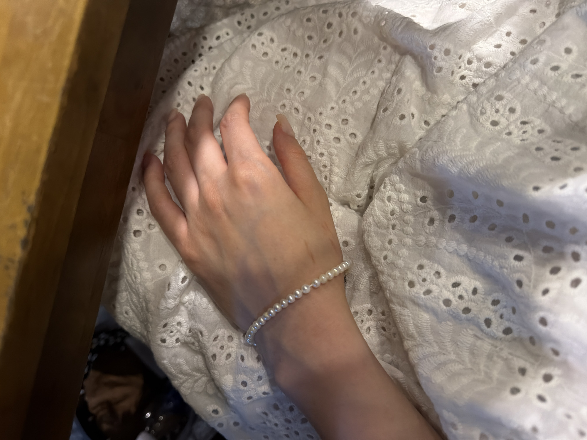 A close-up photograph depicts a hand wearing a pearl bracelet resting on a white eyelet-textured fabric. The hand's fingers are extended. A portion of a wooden surface is visible on the left. The location is not determinable from the provided image.